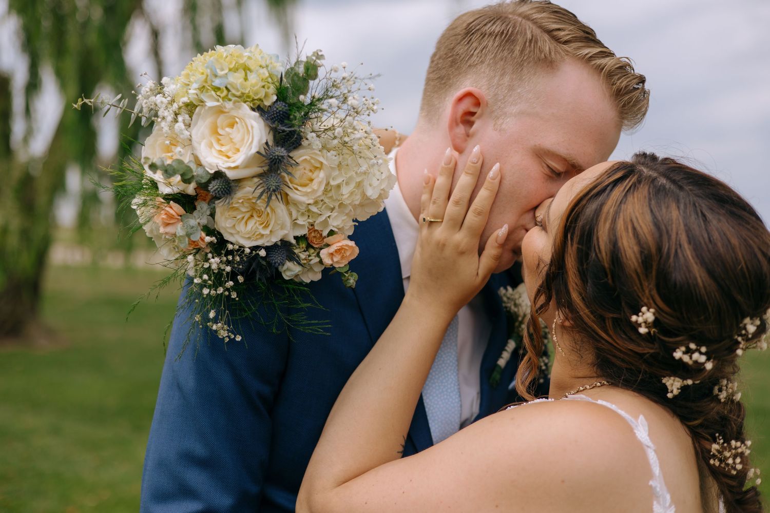 A bride and groom share a romantic kiss while she holds a beautiful white and peach floral bouquet outdoors.