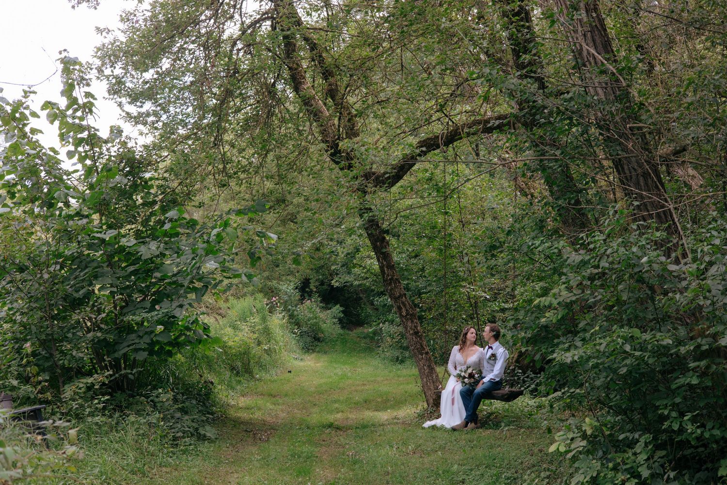 Bride and groom sitting together under a large tree on a scenic green woodland path on their wedding day.