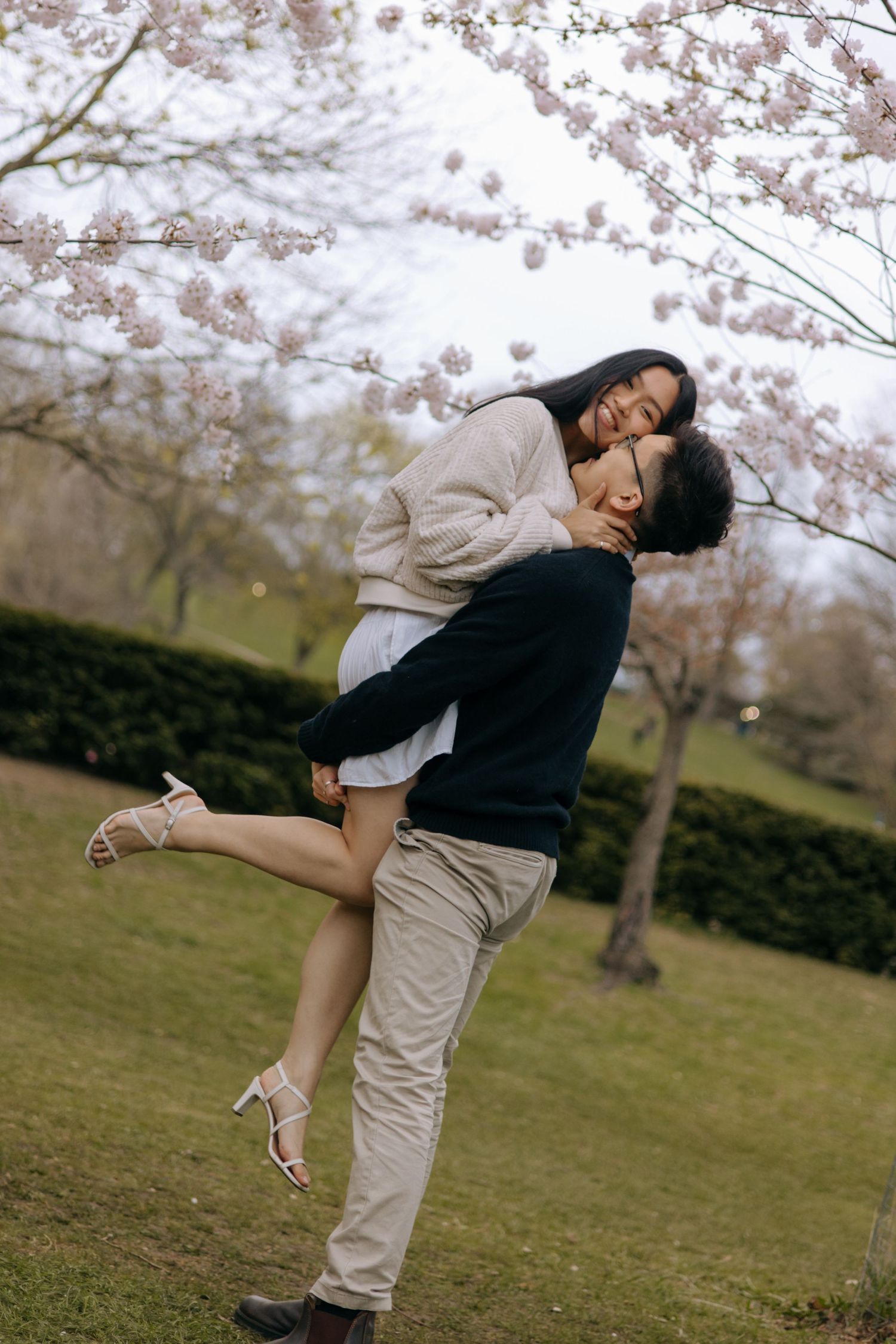 Joyful couple under cherry blossoms as man lifts woman who wraps arms around him laughing.