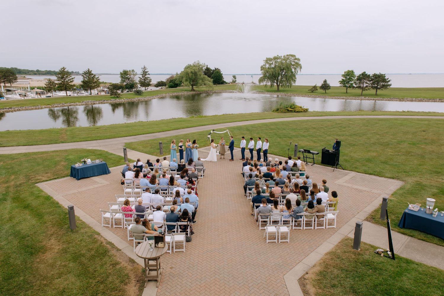 Aerial view of outdoor lakeside wedding ceremony with guests in white chairs on brick patio.