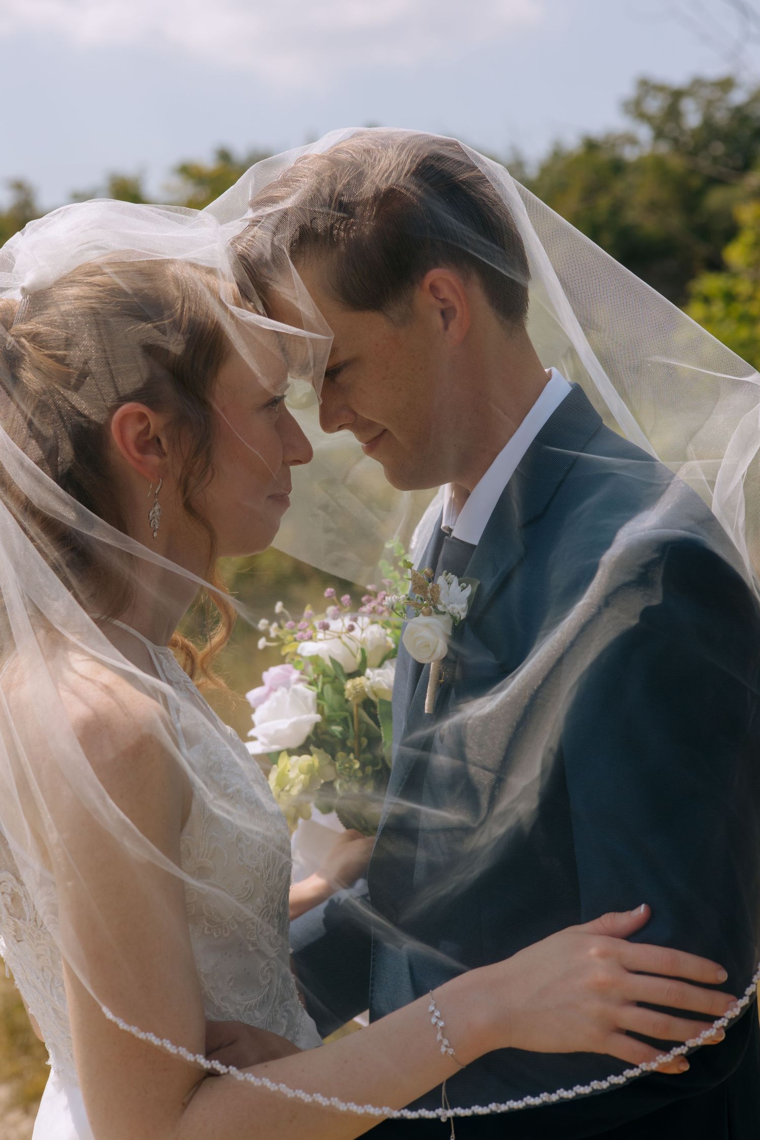 Bride and groom sharing an intimate moment under a flowing white veil outdoors on a sunny day.