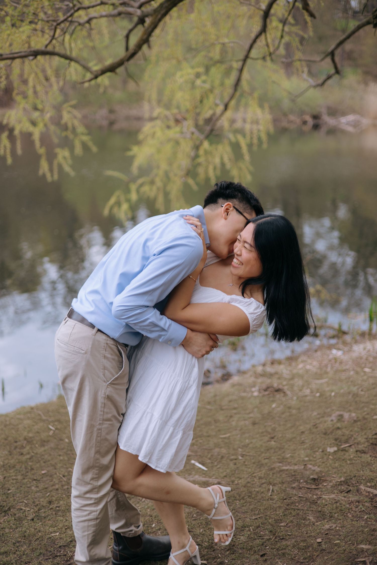 Joyful Asian couple embracing and laughing together beside a tranquil pond under willow trees.