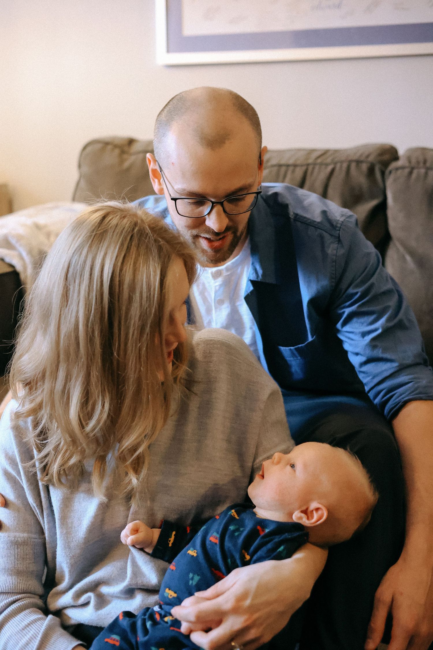 Smiling couple with glasses sitting on a couch together, lovingly looking at their baby.