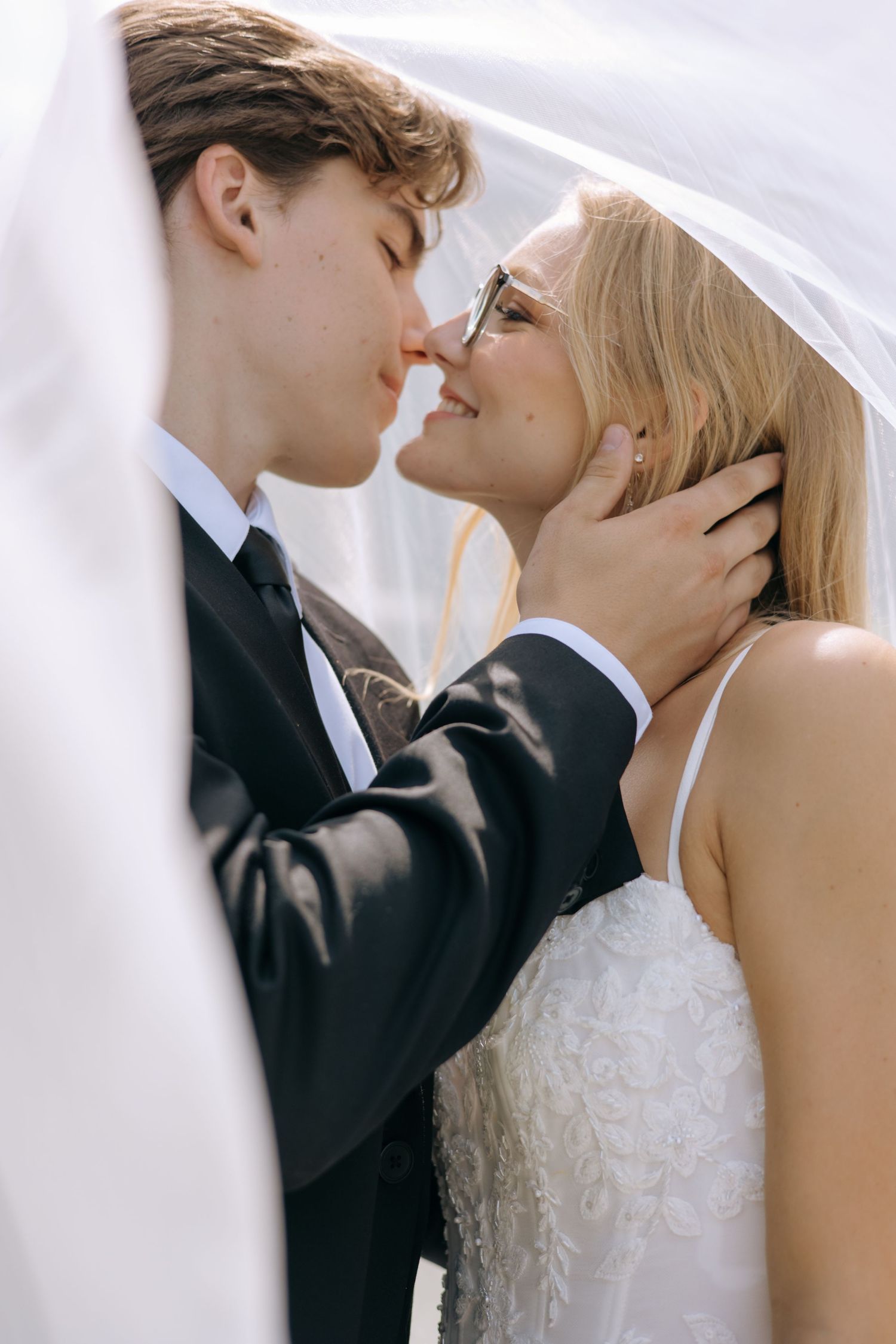 A bride and groom share a romantic moment under a white veil, nearly kissing on their wedding day.
