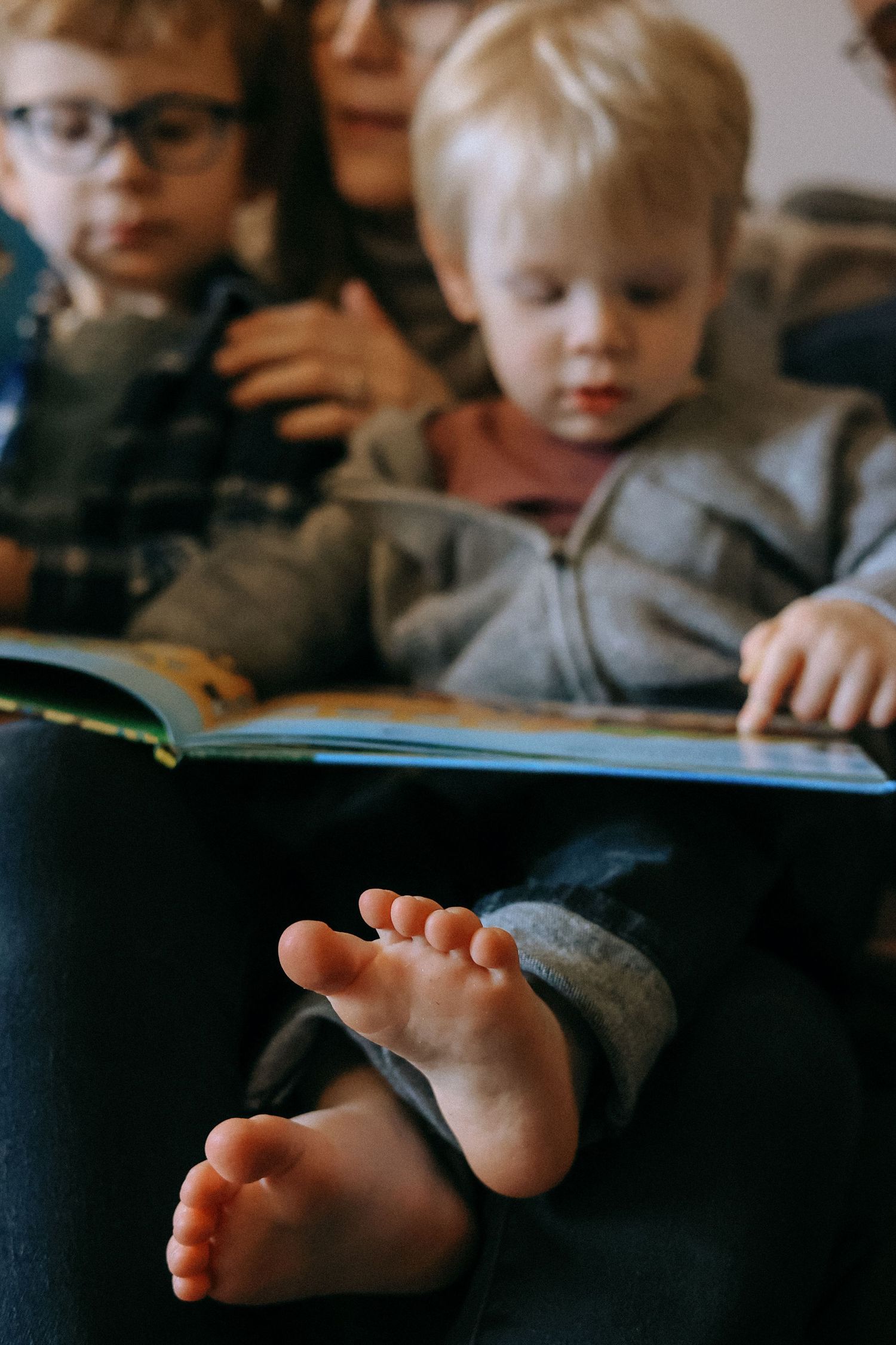 Young children reading a colorful picture book together with tiny bare baby feet in foreground.