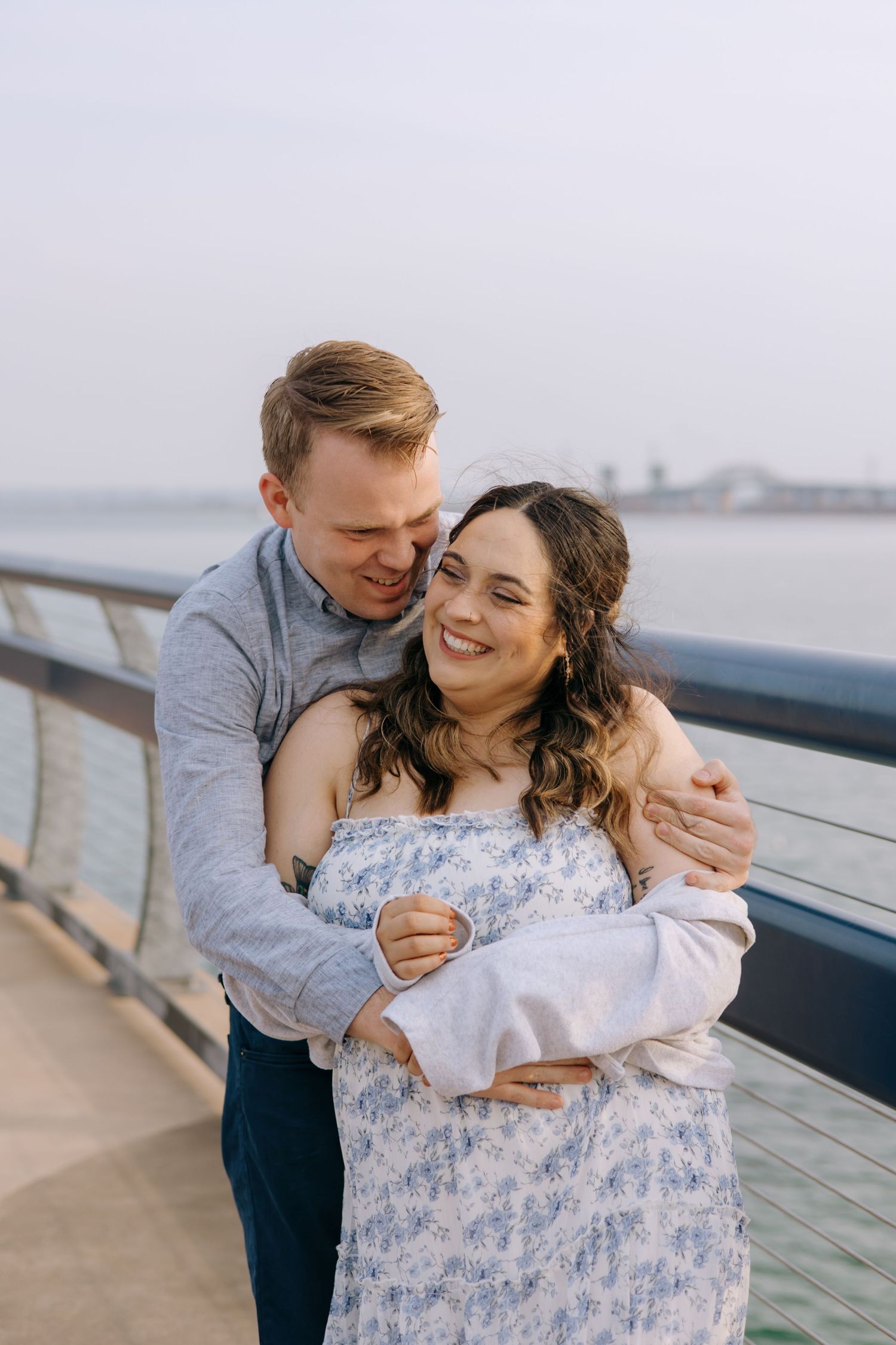 Happy couple embracing on a waterfront pier, woman in floral dress smiling as man hugs her from behind.