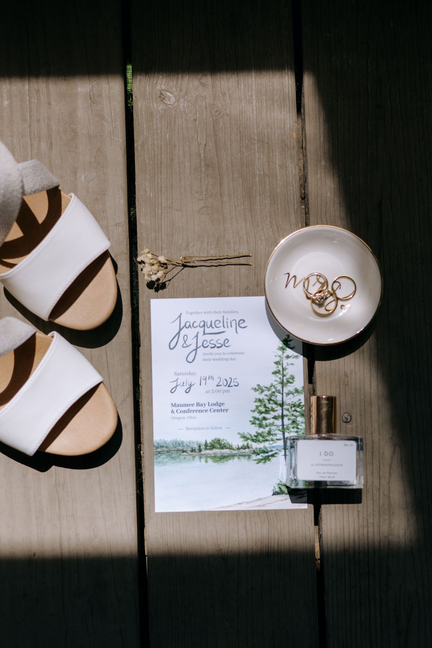 Wedding flat lay featuring white sandals, floral invitation, gold rings dish, and perfume on wood.