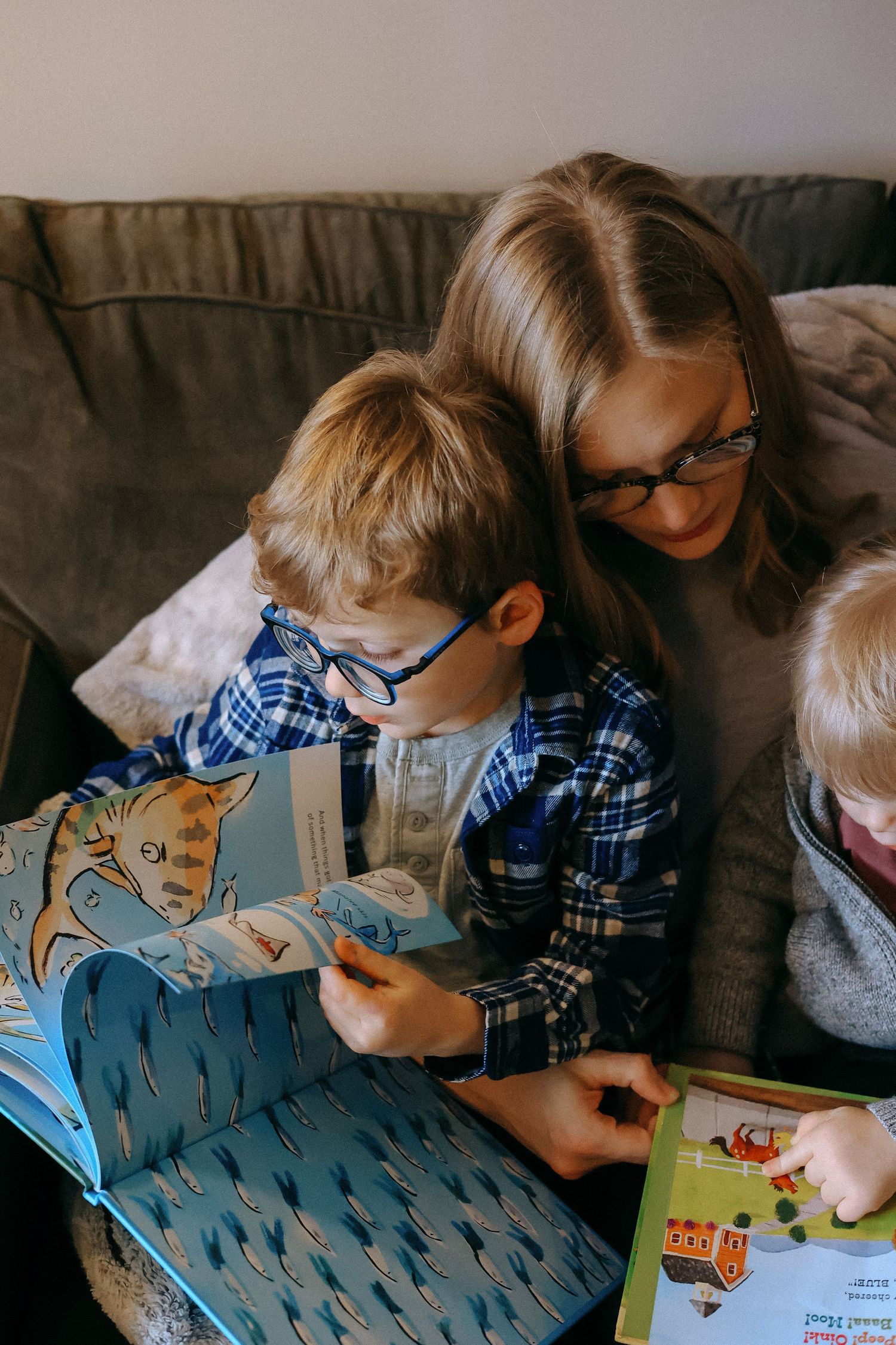 Mother with glasses reads colorful picture books with two young boys on a cozy couch.