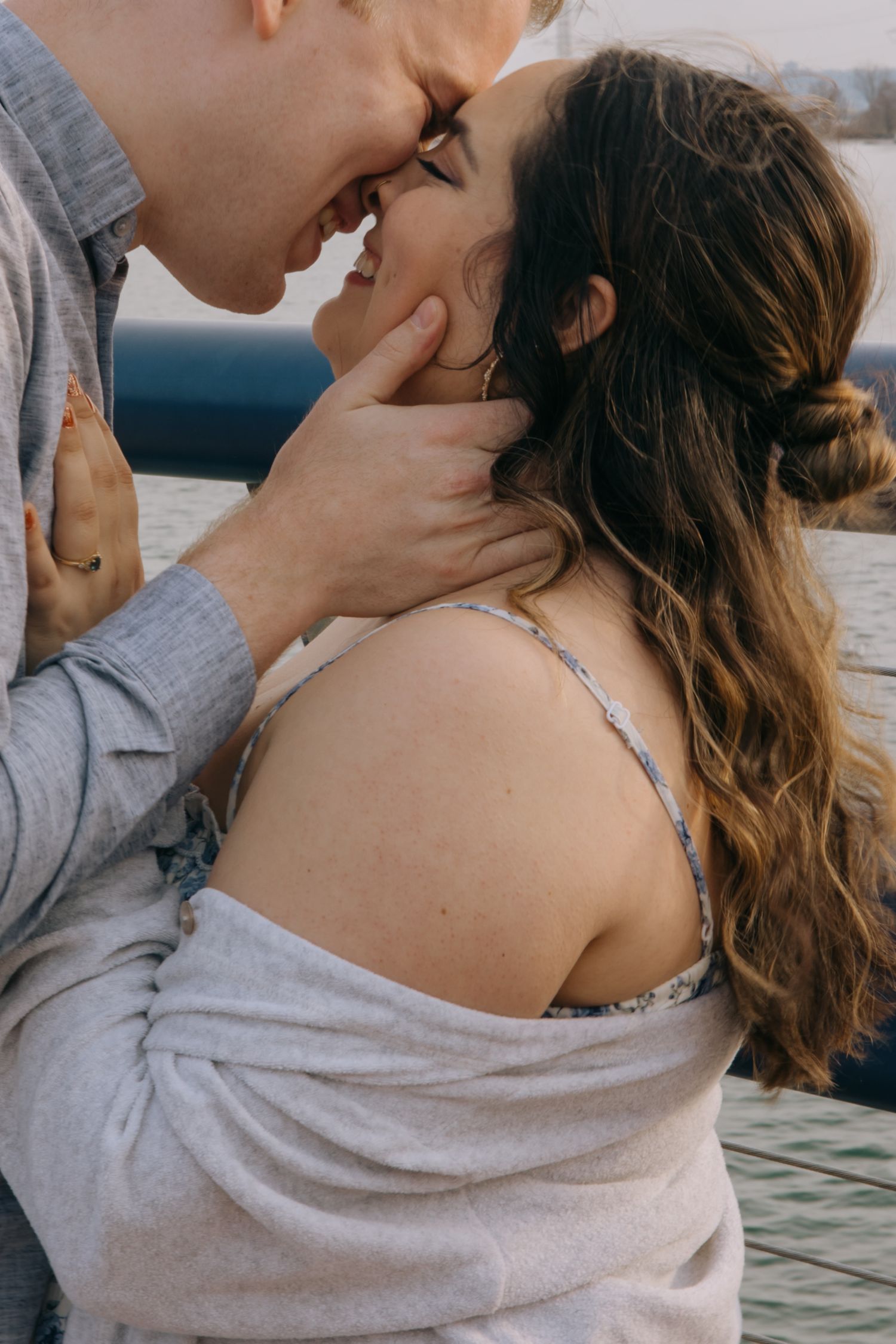 Intimate close-up of couple almost kissing outdoors, man tenderly holding womans face near waterfront.