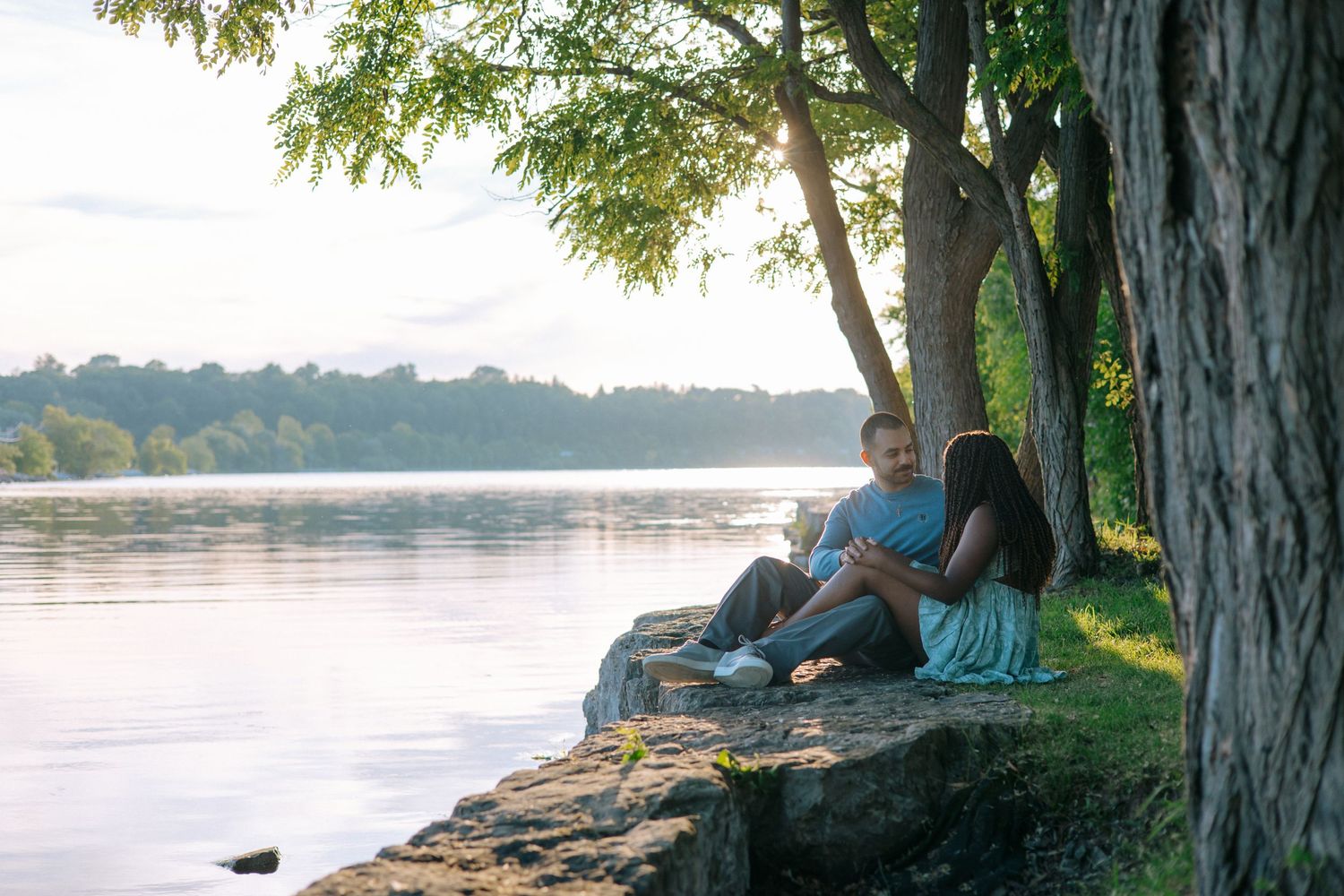 Couple sitting together on rocky shoreline under trees at sunset by a calm river.