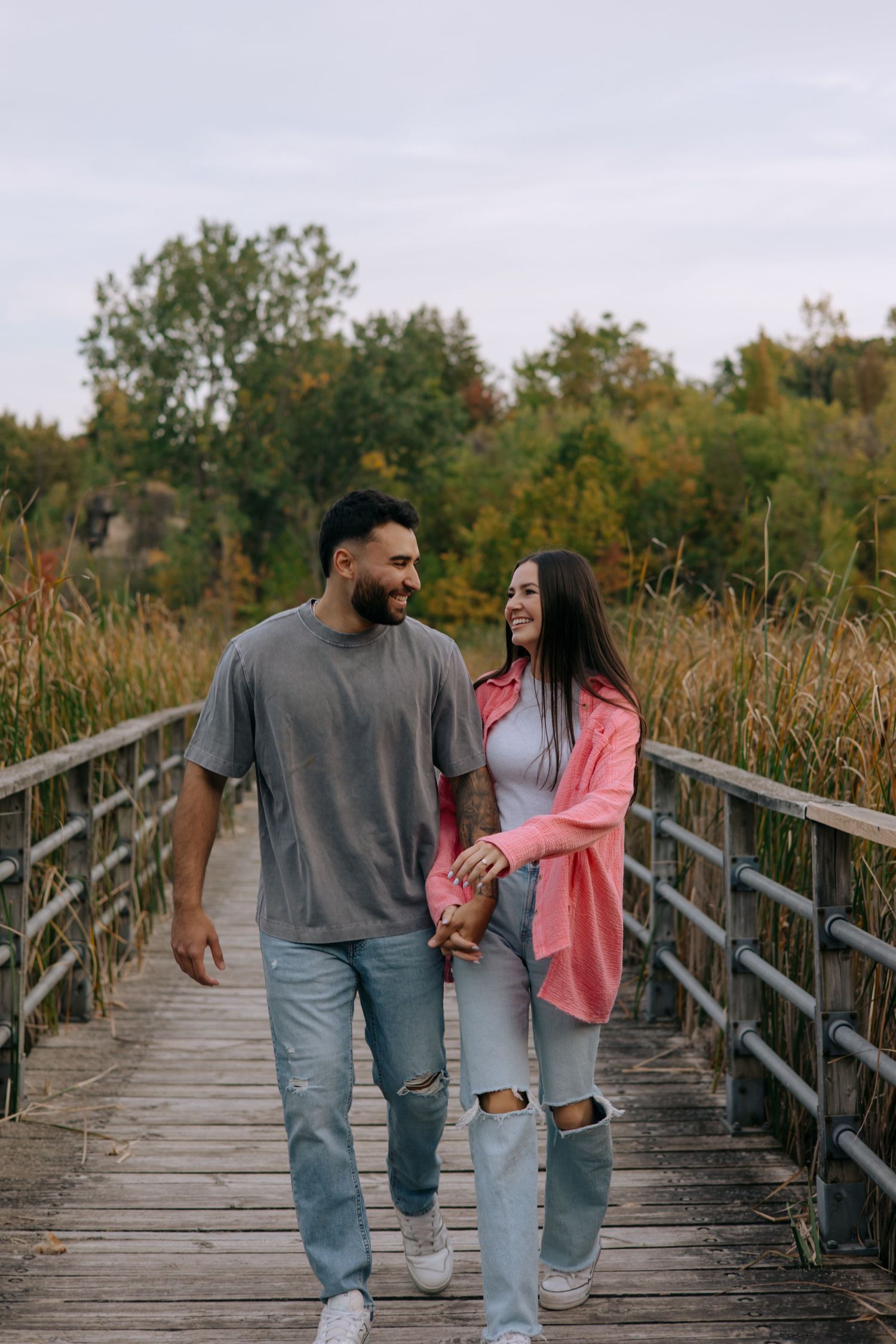 Smiling couple holding hands while walking on a wooden bridge surrounded by autumn foliage.