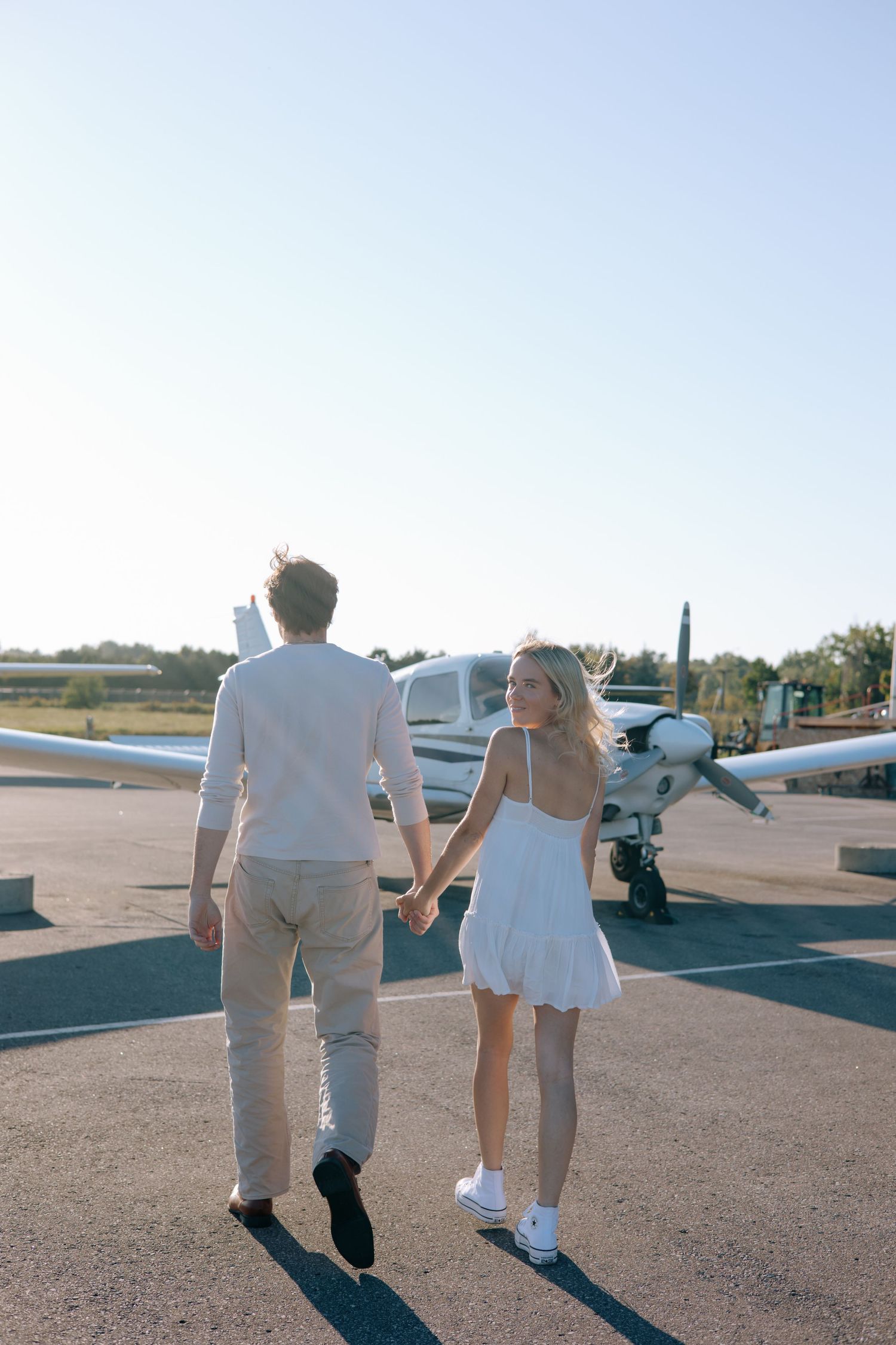 Couple walking hand in hand toward a small white propeller plane on a sunny airport tarmac.