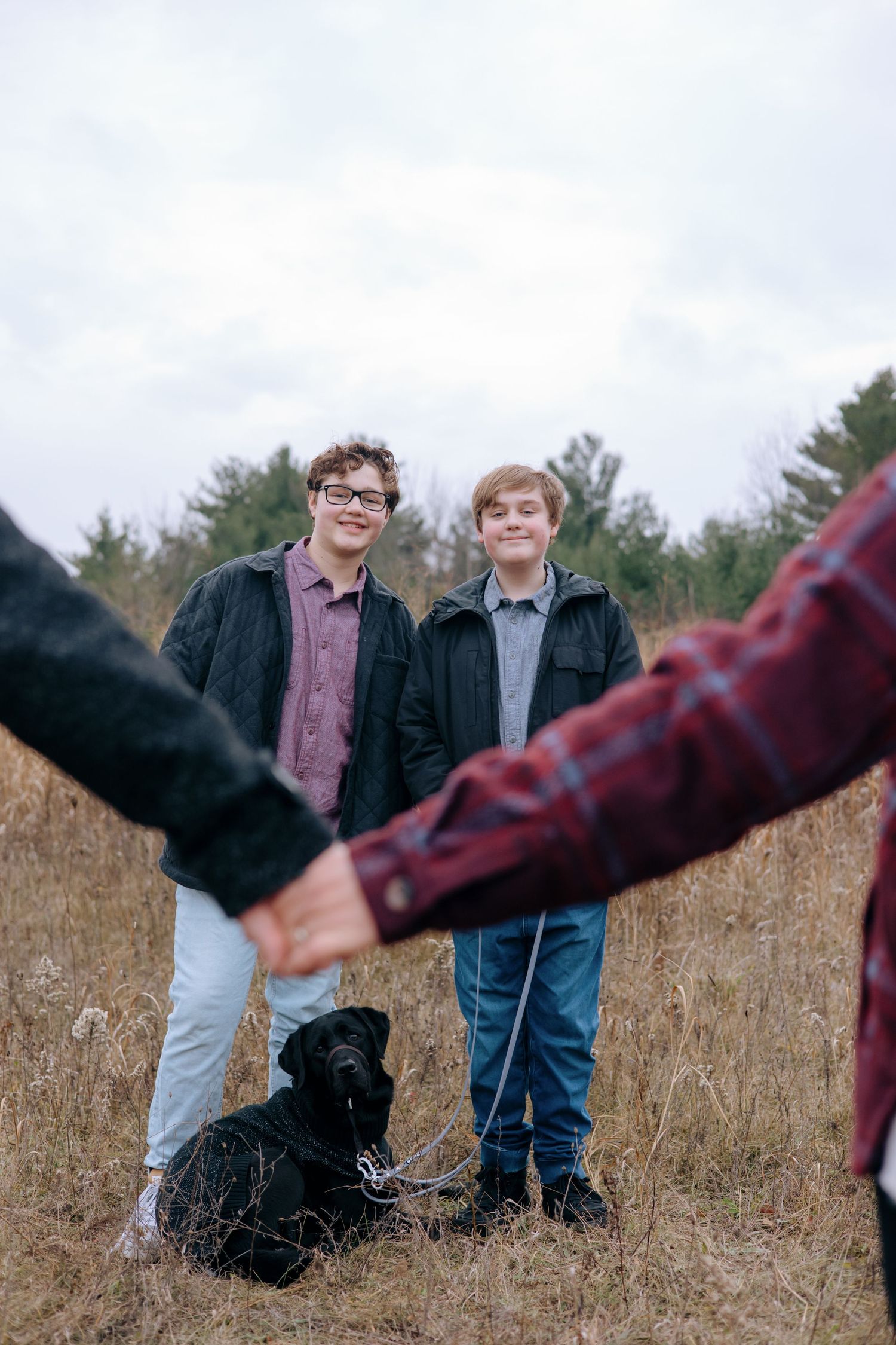 Two teenage boys smiling in autumn field with black Labrador dog, parents holding hands in foreground.
