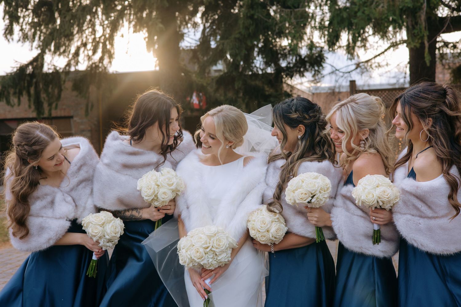 Bride surrounded by bridesmaids in navy dresses and white fur shawls holding white floral bouquets outdoors.