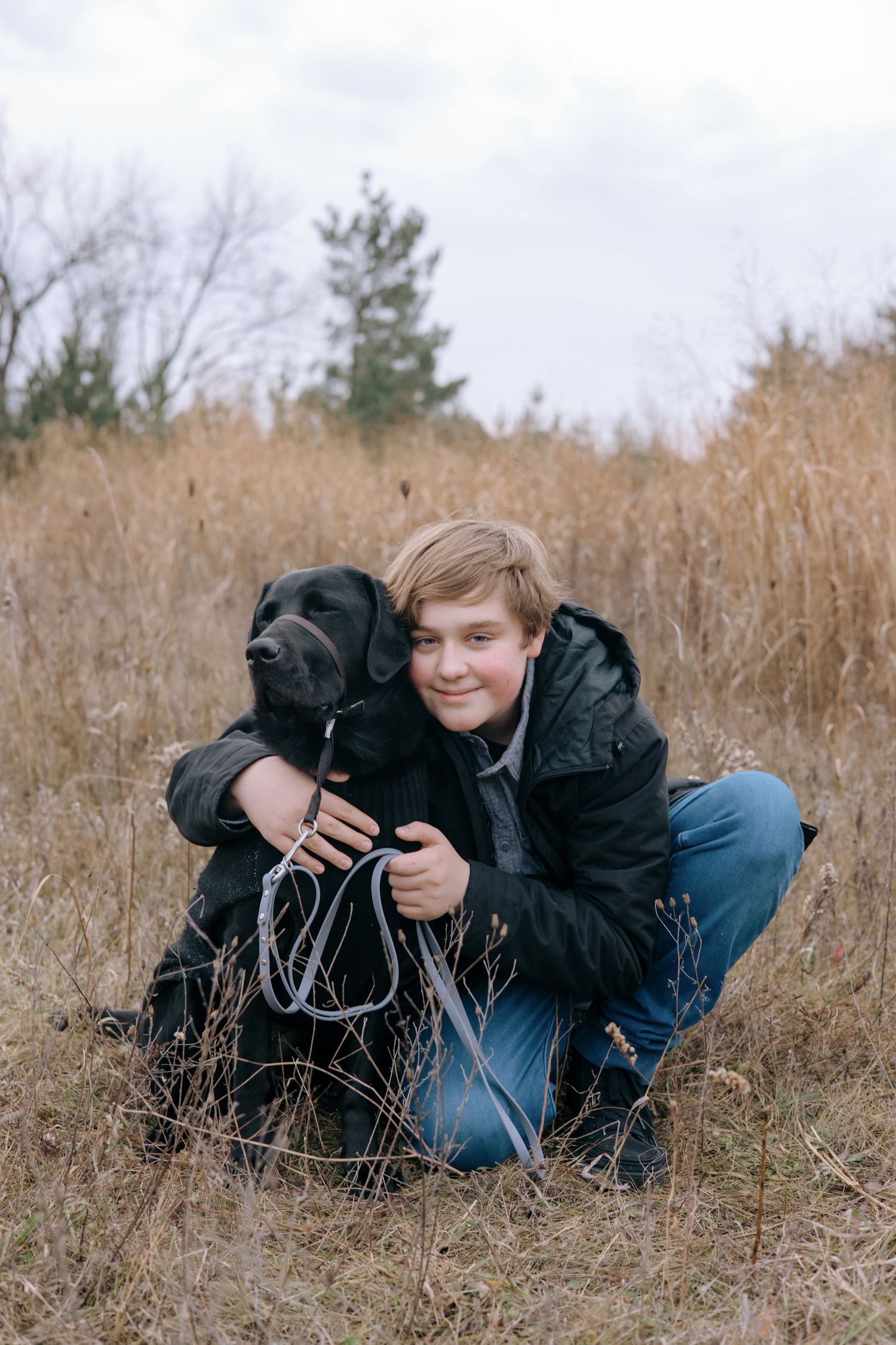 Smiling young boy crouching in dry grass field hugging black Labrador dog on a cloudy autumn day.