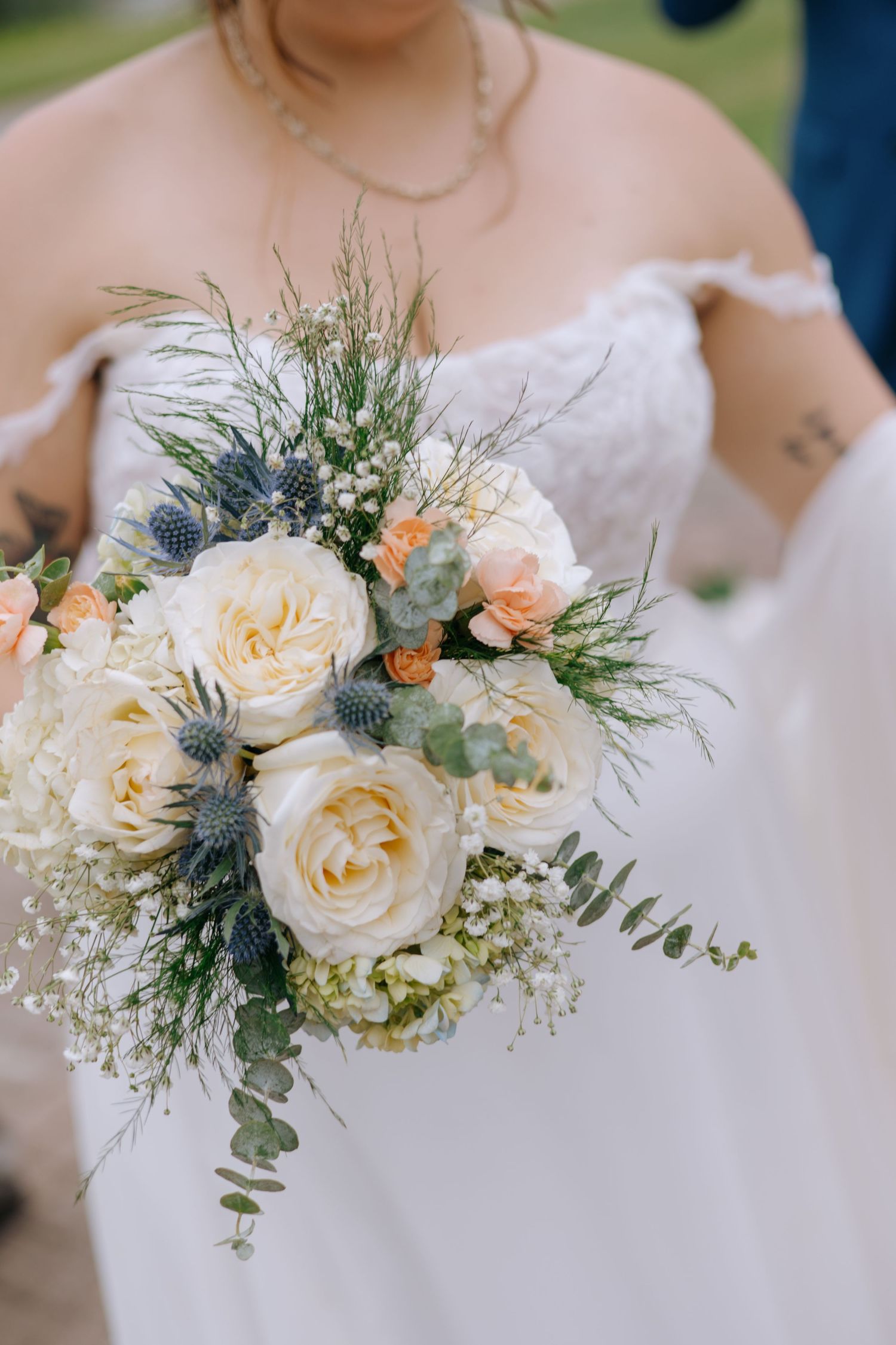 Bride holding elegant wedding bouquet with white roses, peach blooms, blue thistle, and eucalyptus in white lace gown.