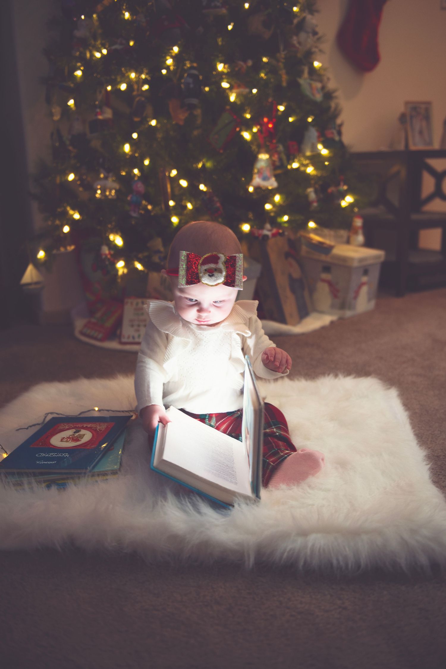 Child lying on white fur rug reading illuminated book near Christmas tree.