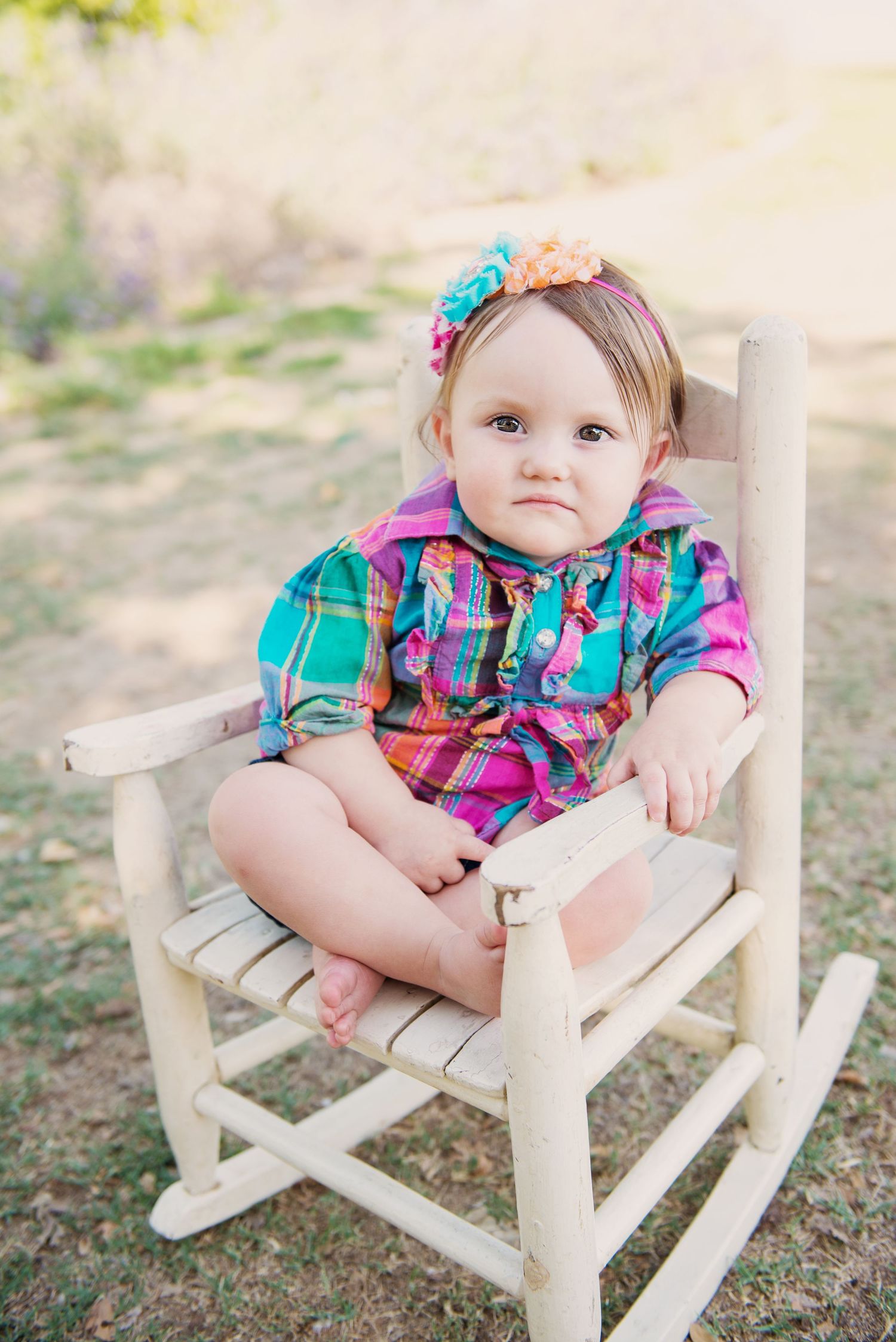 Baby in plaid outfit and floral headband sitting in white rocking chair.