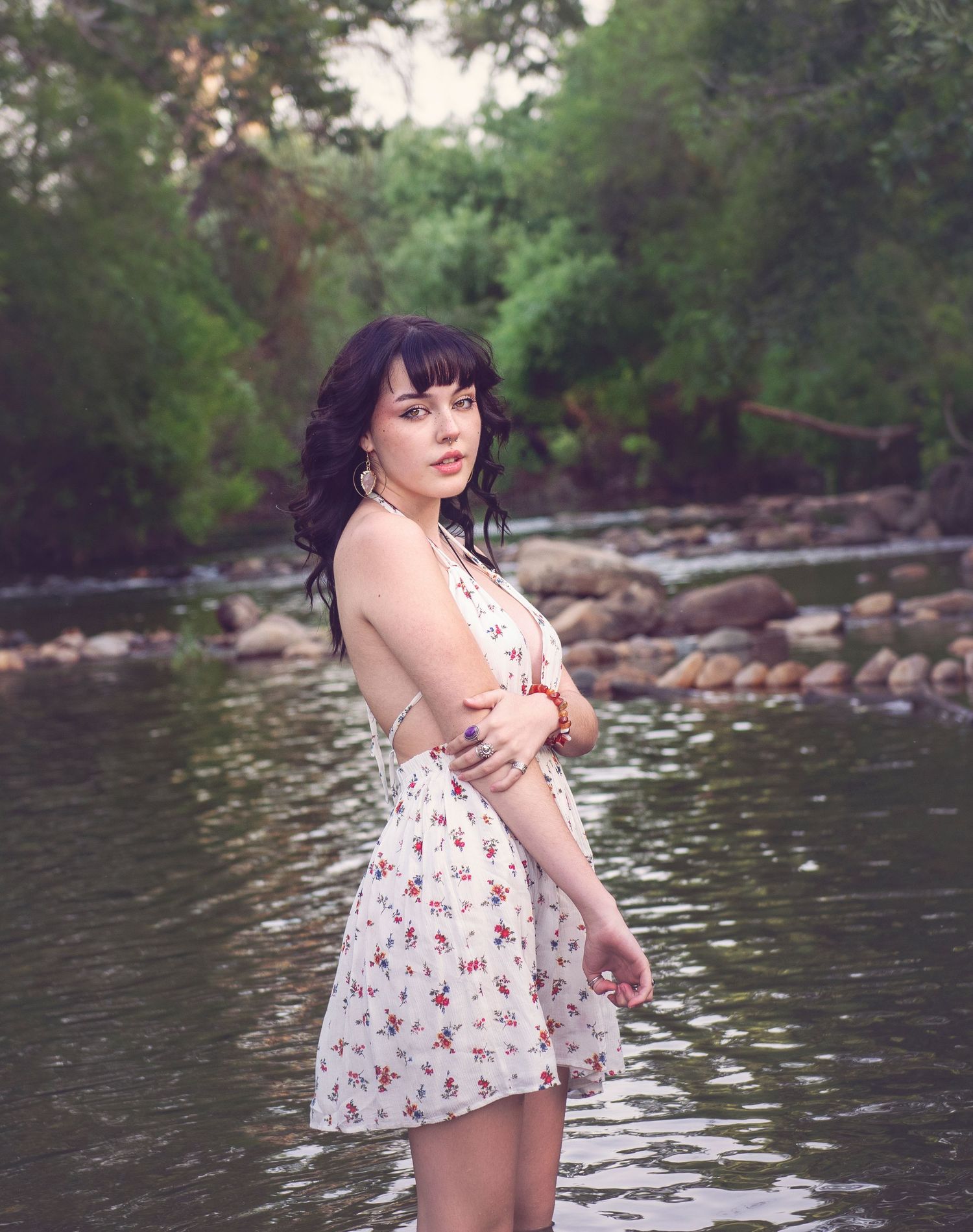 Figure in floral dress poses beside flowing river with rocky shoreline.