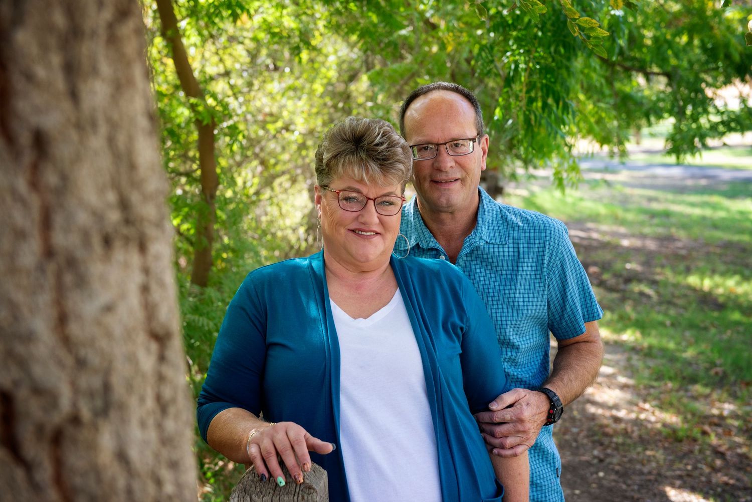 Couple poses for photos outdoors under green leafy trees on sunny day.