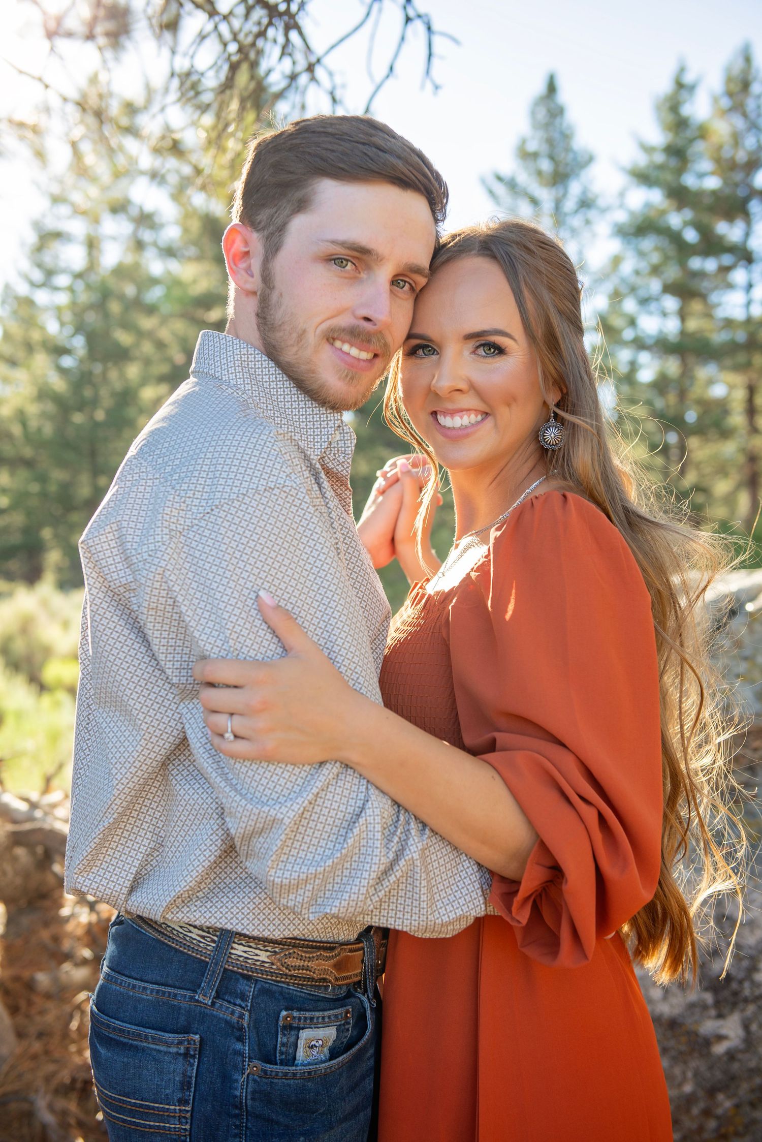 A couple embraces during their engagement photoshoot in a sunny forest setting.
