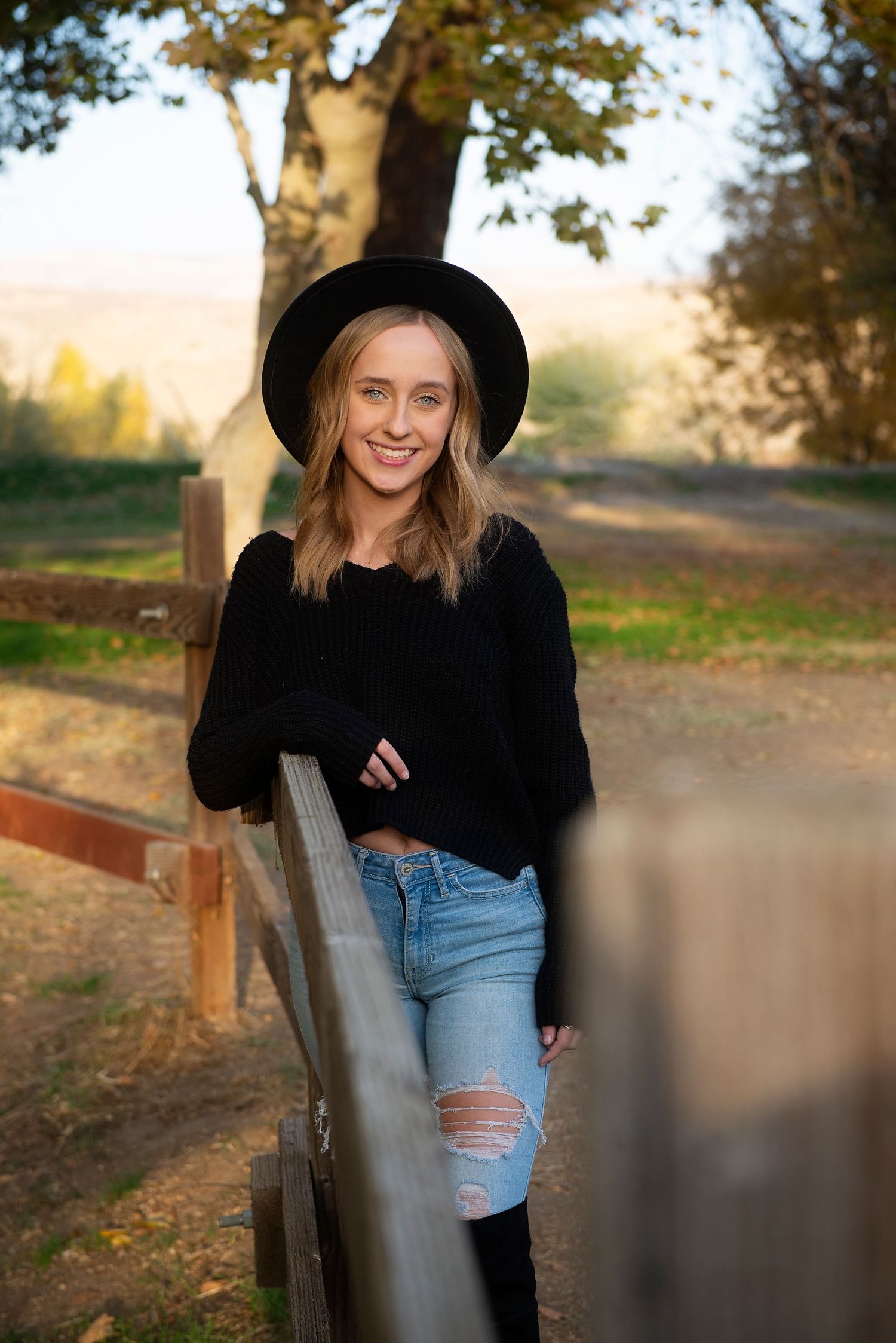 Person wearing black sweater and ripped jeans poses against wooden fence in autumn scenery.