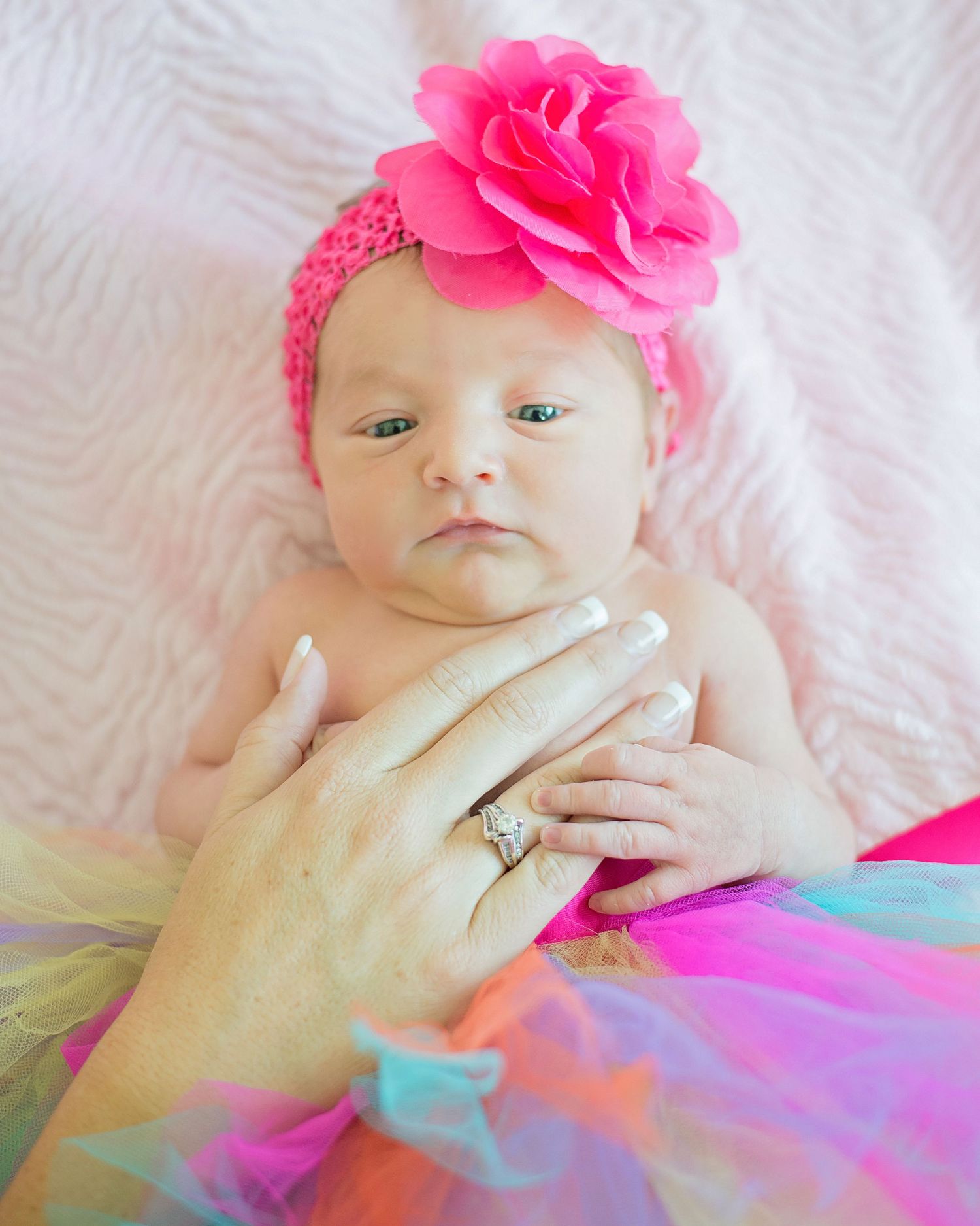 Baby wearing bright pink flower headband and colorful tutu lying on white textured fabric.