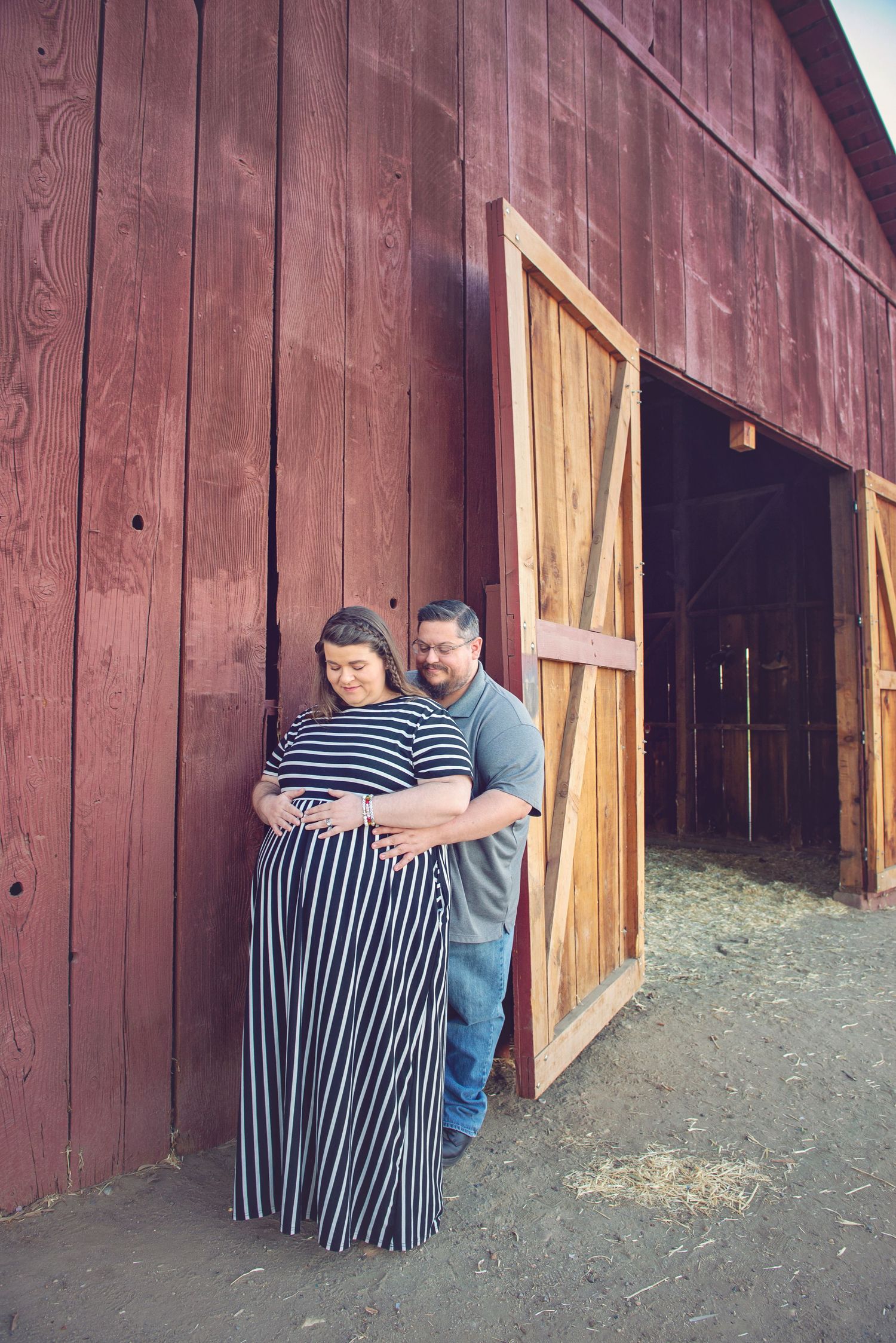 Couple poses together against red barn door in matching striped outfits. Maternity