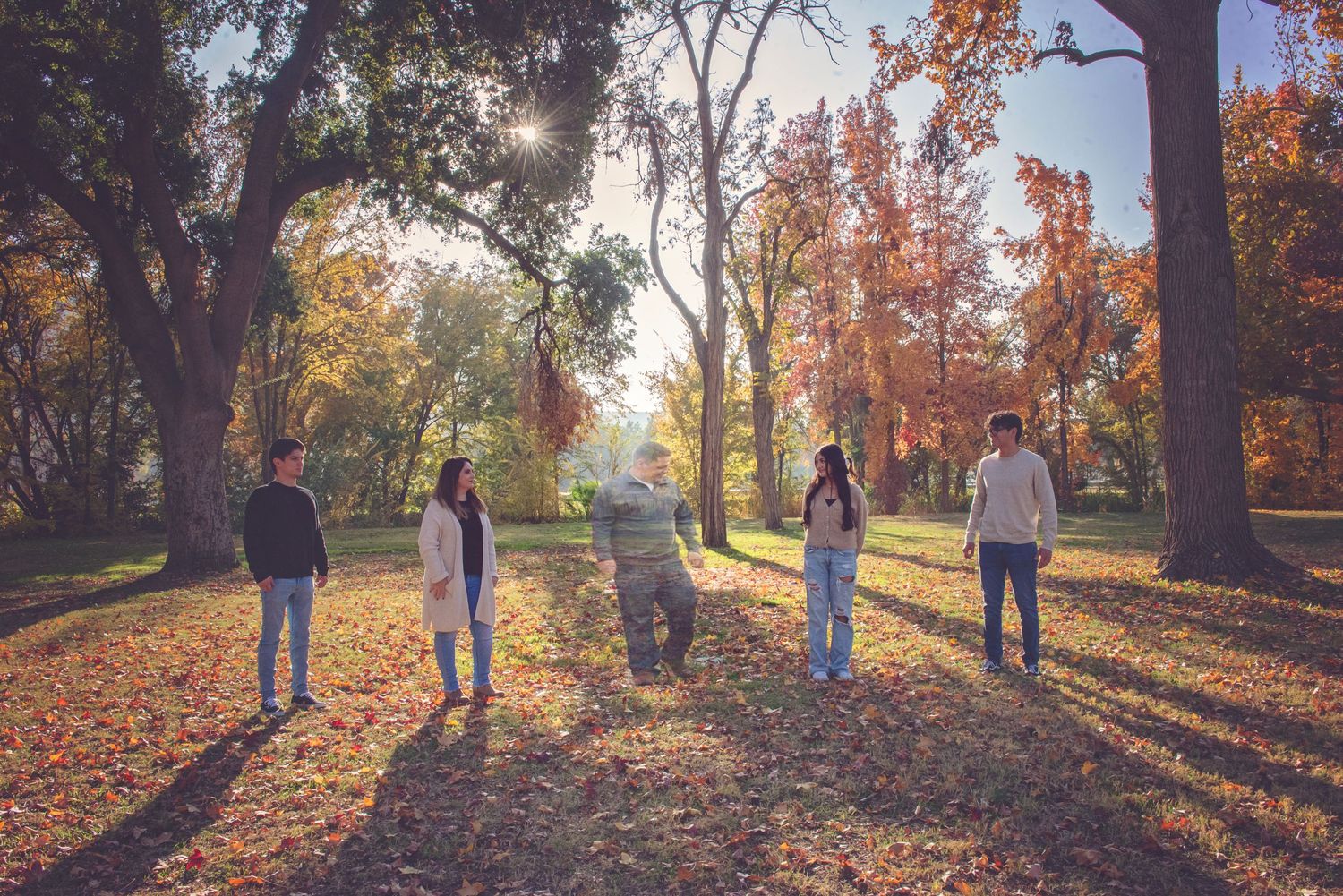 Family walking through park with golden autumn leaves and sunlight filtering through trees.