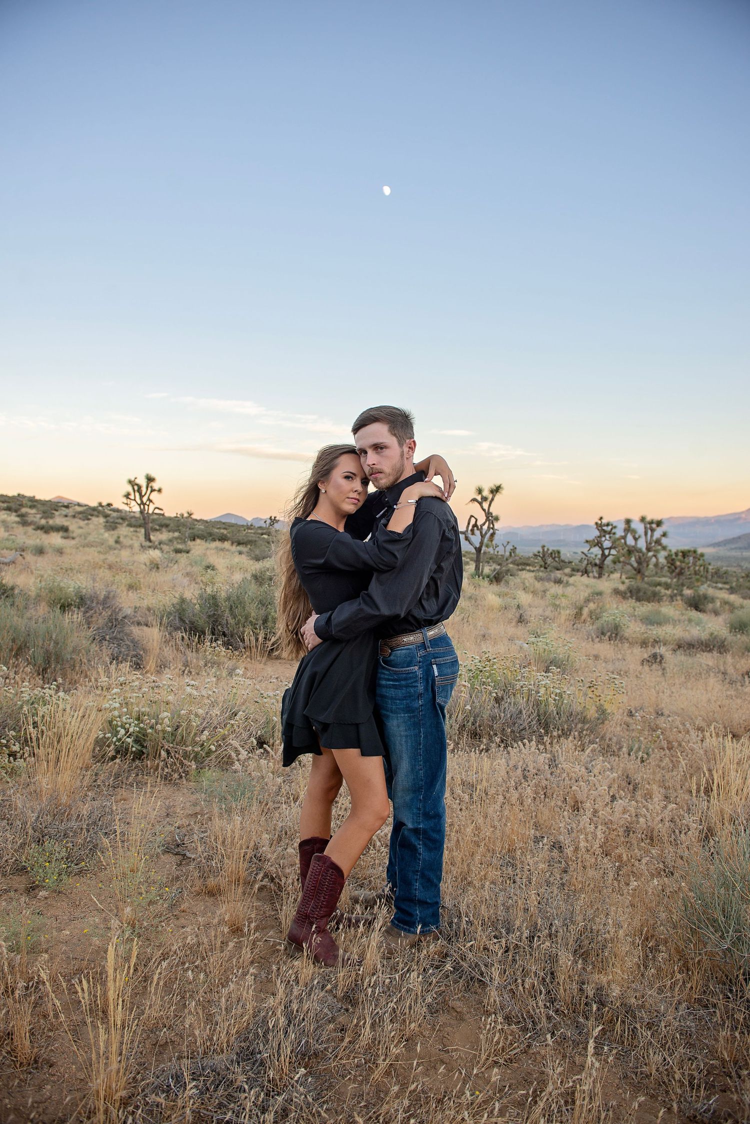 A couple embraces in a desert landscape with Joshua trees and mountains at golden hour.