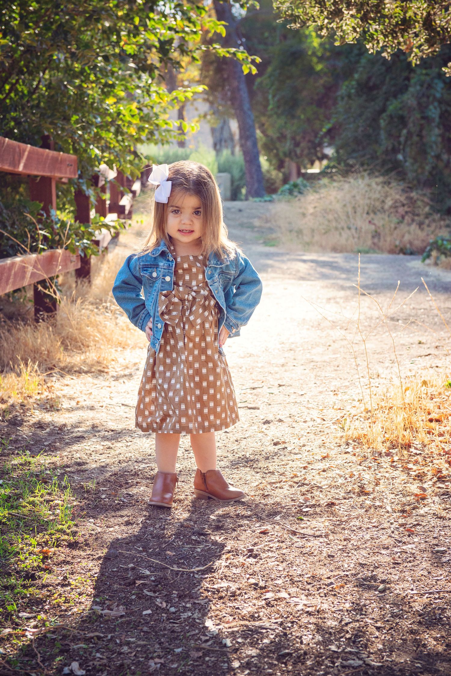 Small toddler in a floral dress and denim jacket walks along a sunlit autumn path.