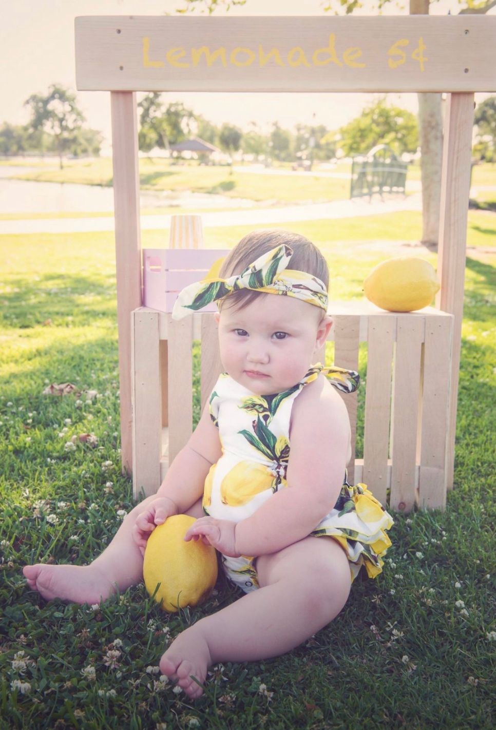 Baby in yellow outfit poses with lemons next to a white wooden fence in summer.
