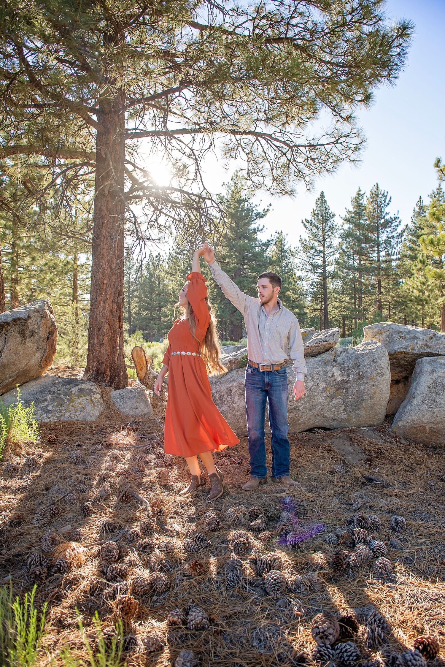 A couple celebrates on rocky terrain surrounded by pine trees in warm sunlight.