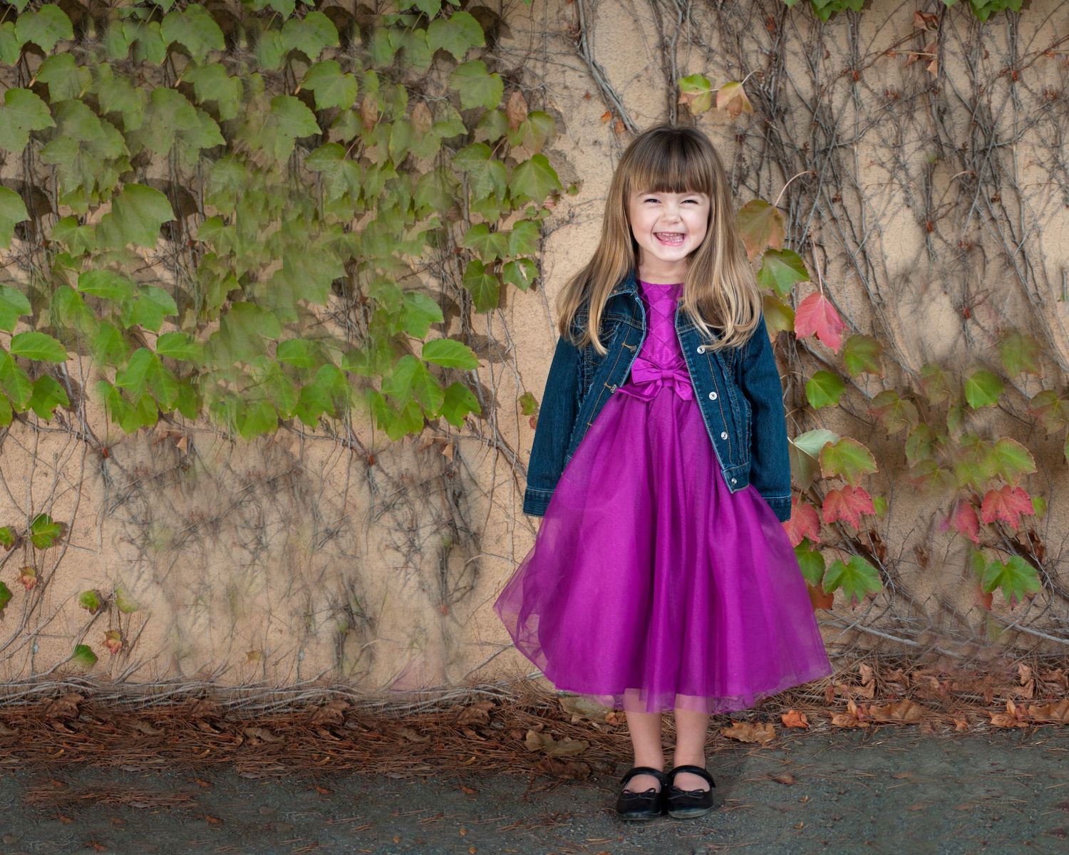 Child in vibrant purple dress twirling against textured stone wall with climbing vines.