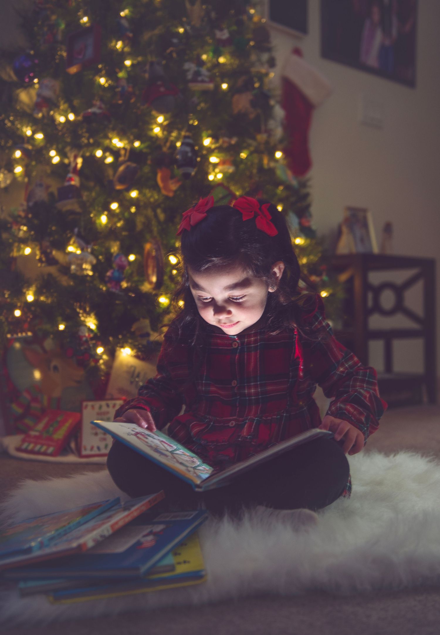 Child reading book by Christmas tree with glowing lights in dark room.