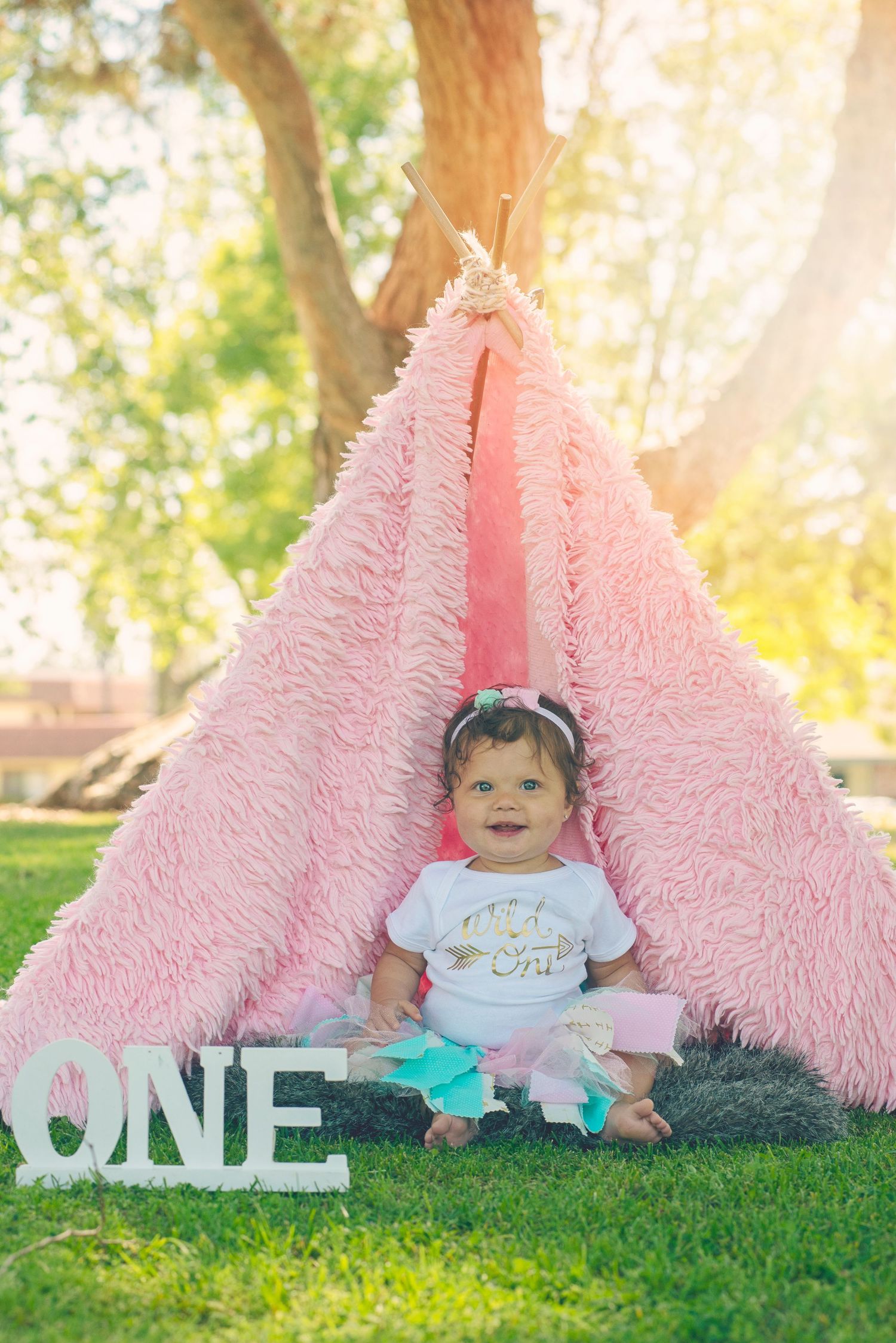 Baby sits by pink fuzzy teepee tent with 'ONE' letters during milestone photo session.