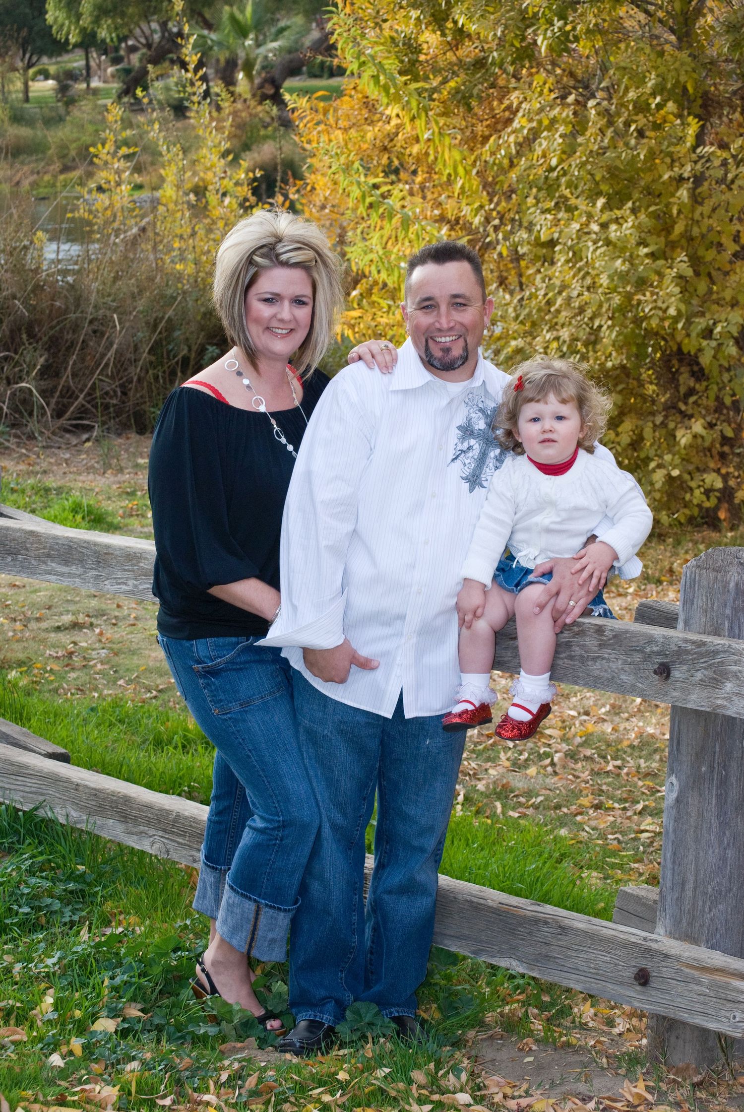 Family group photo by wooden fence with autumn foliage in background.