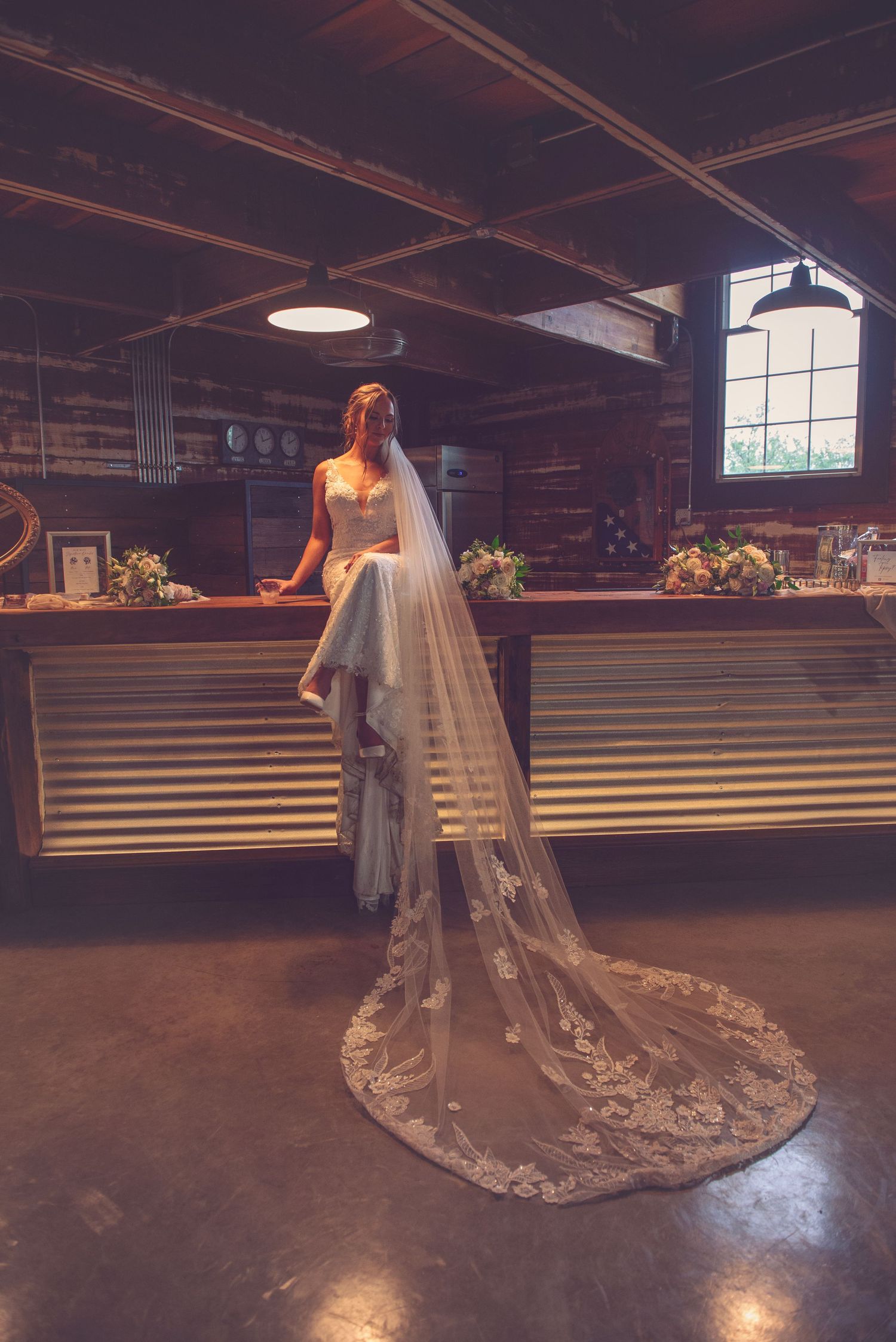 Long lace wedding dress train spreads across rustic wooden floor in indoor venue.