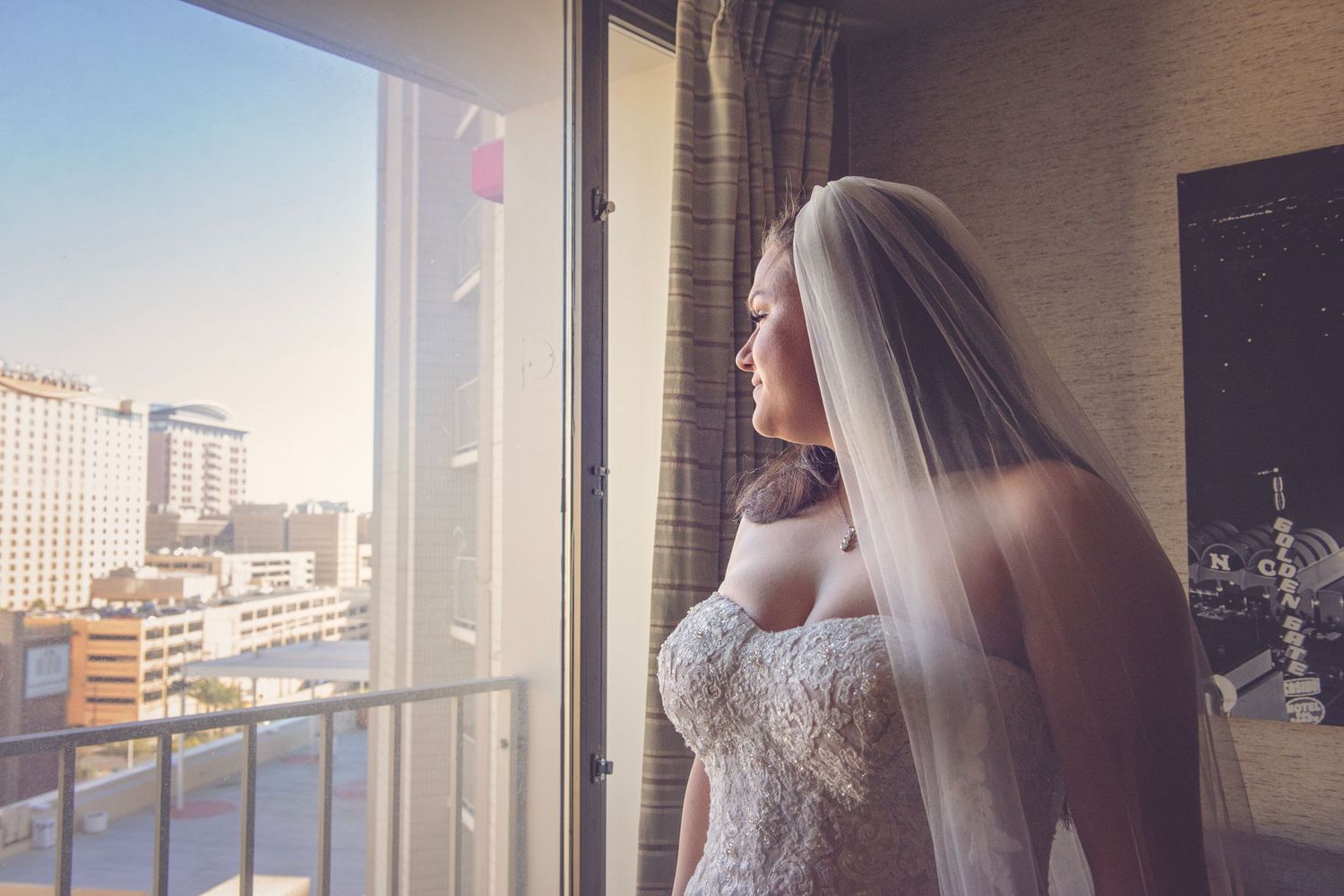 Bride in wedding dress stands near window overlooking city skyline.