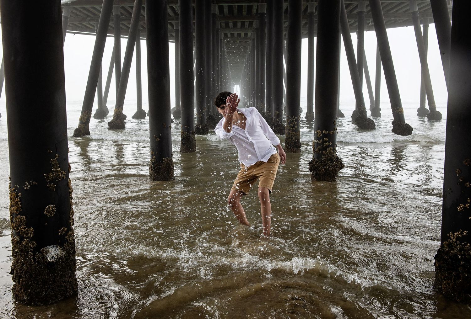 Black and white sequence of figures exploring under a pier with wooden pilings and rippling water.