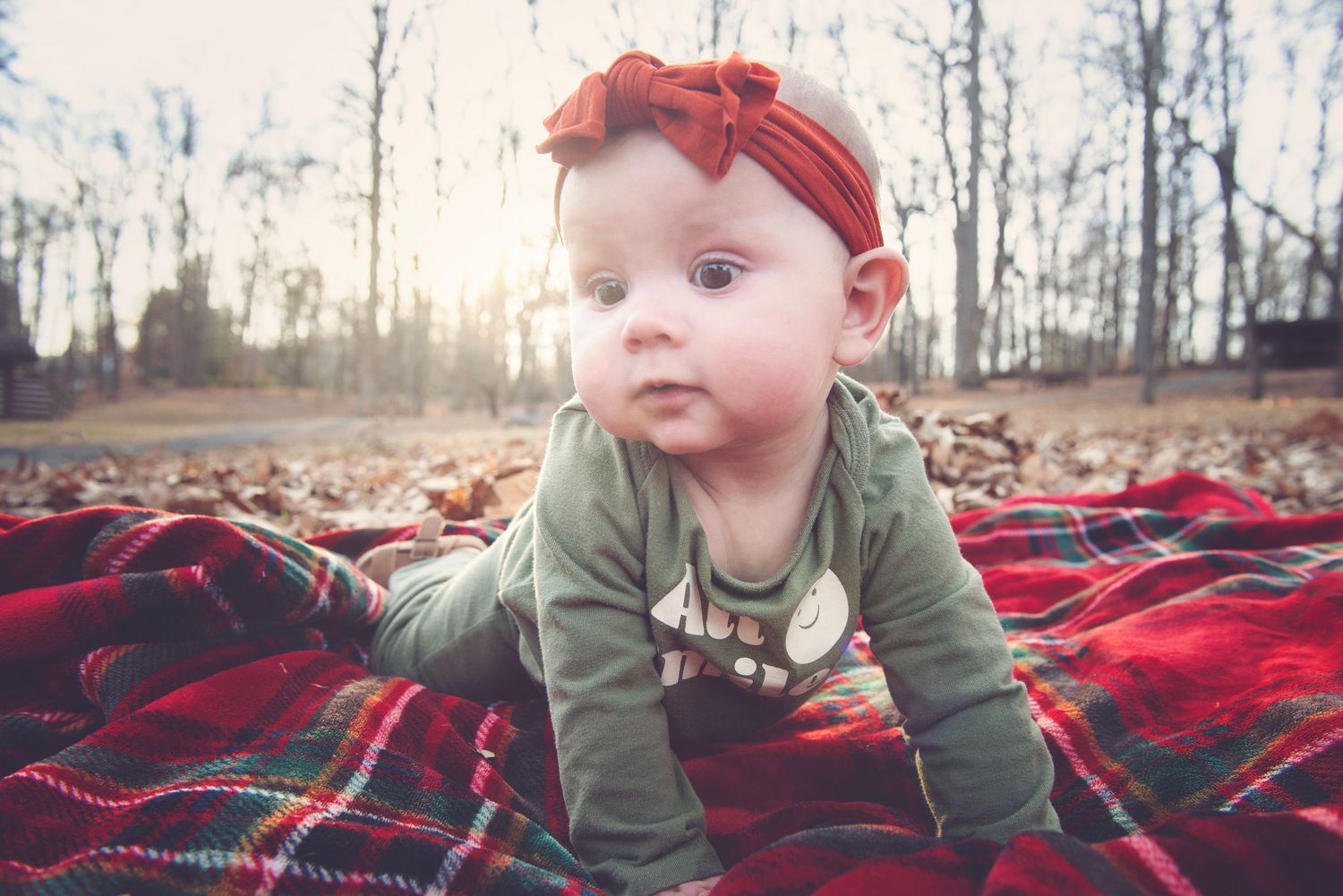 Baby in olive green clothing laying on red plaid blanket during autumn photoshoot.