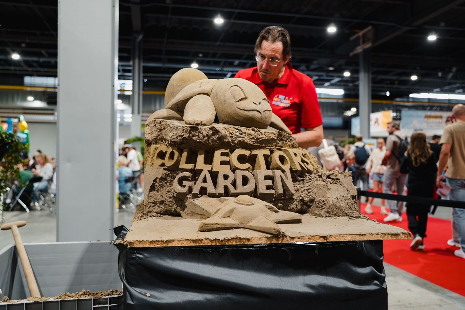 Artist sculpting a detailed sand sculpture reading Collectors Garden at an indoor exhibition event.