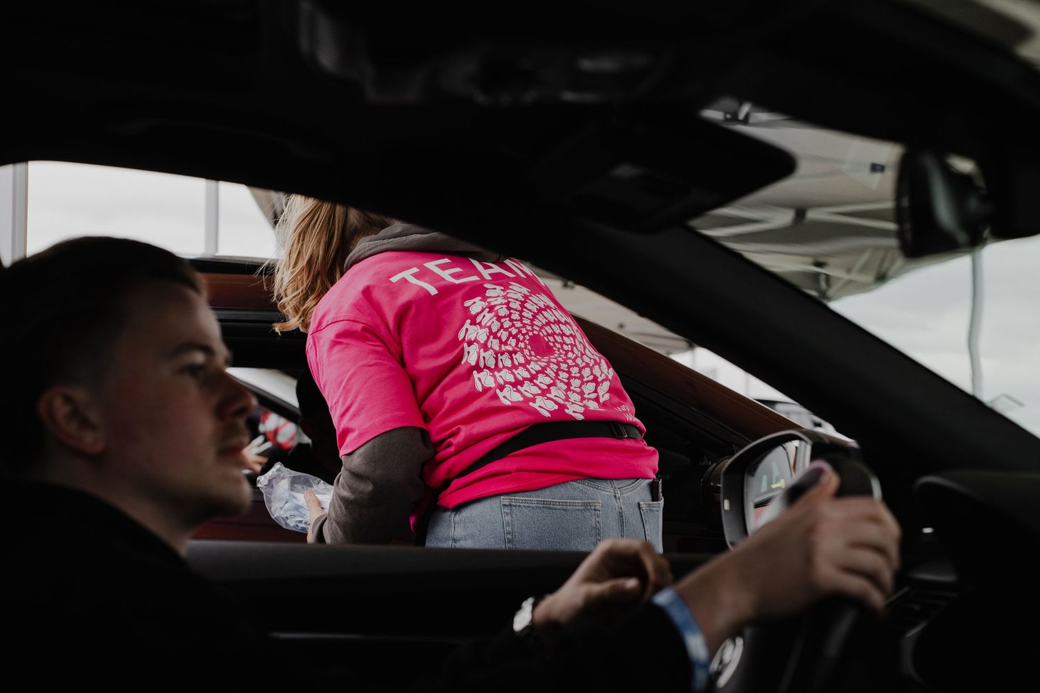 Person in pink TEAM shirt leaning into a car window while driver holds steering wheel at motorsport event.