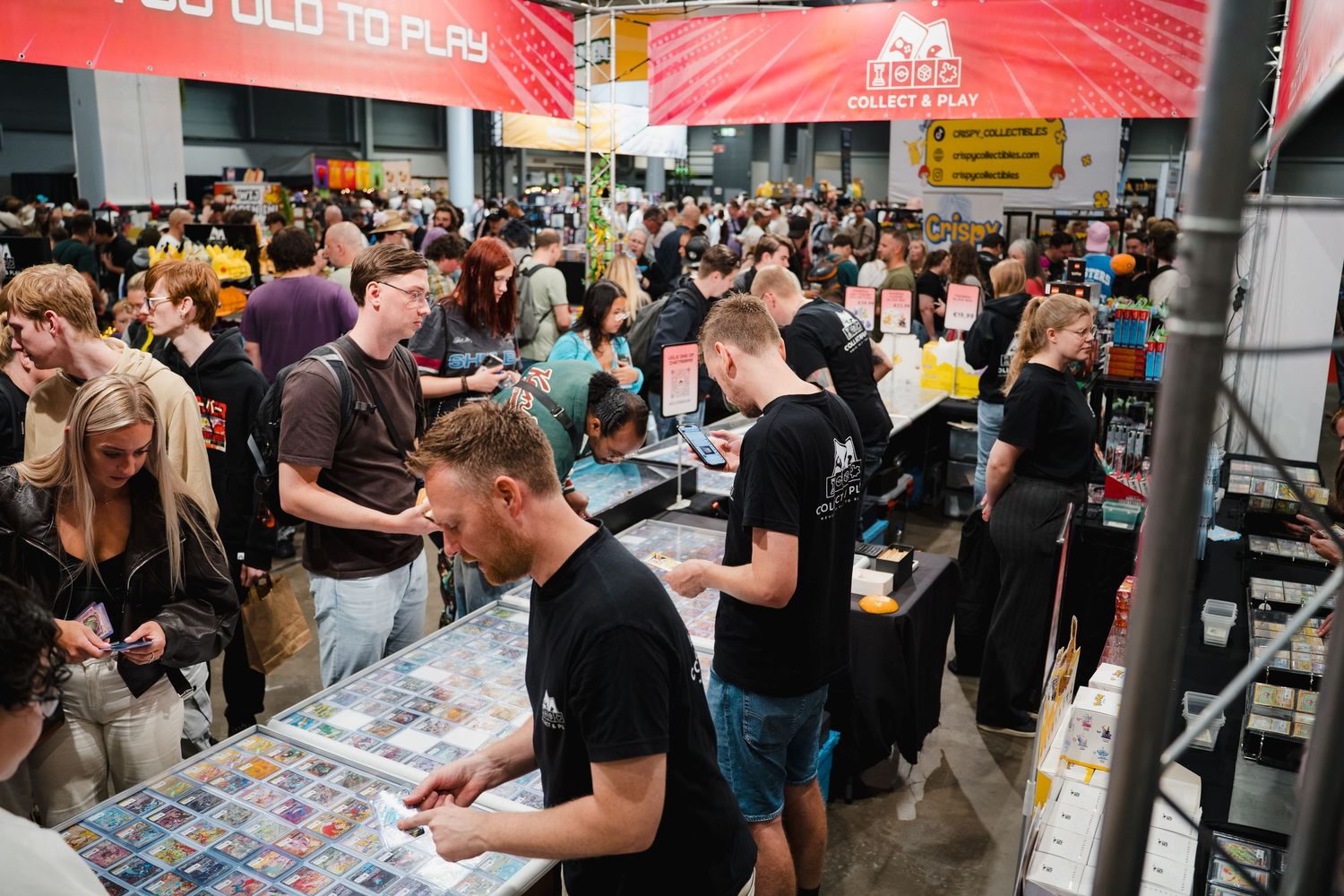 Crowded gaming convention hall with players browsing card game tables under a red promotional banner.