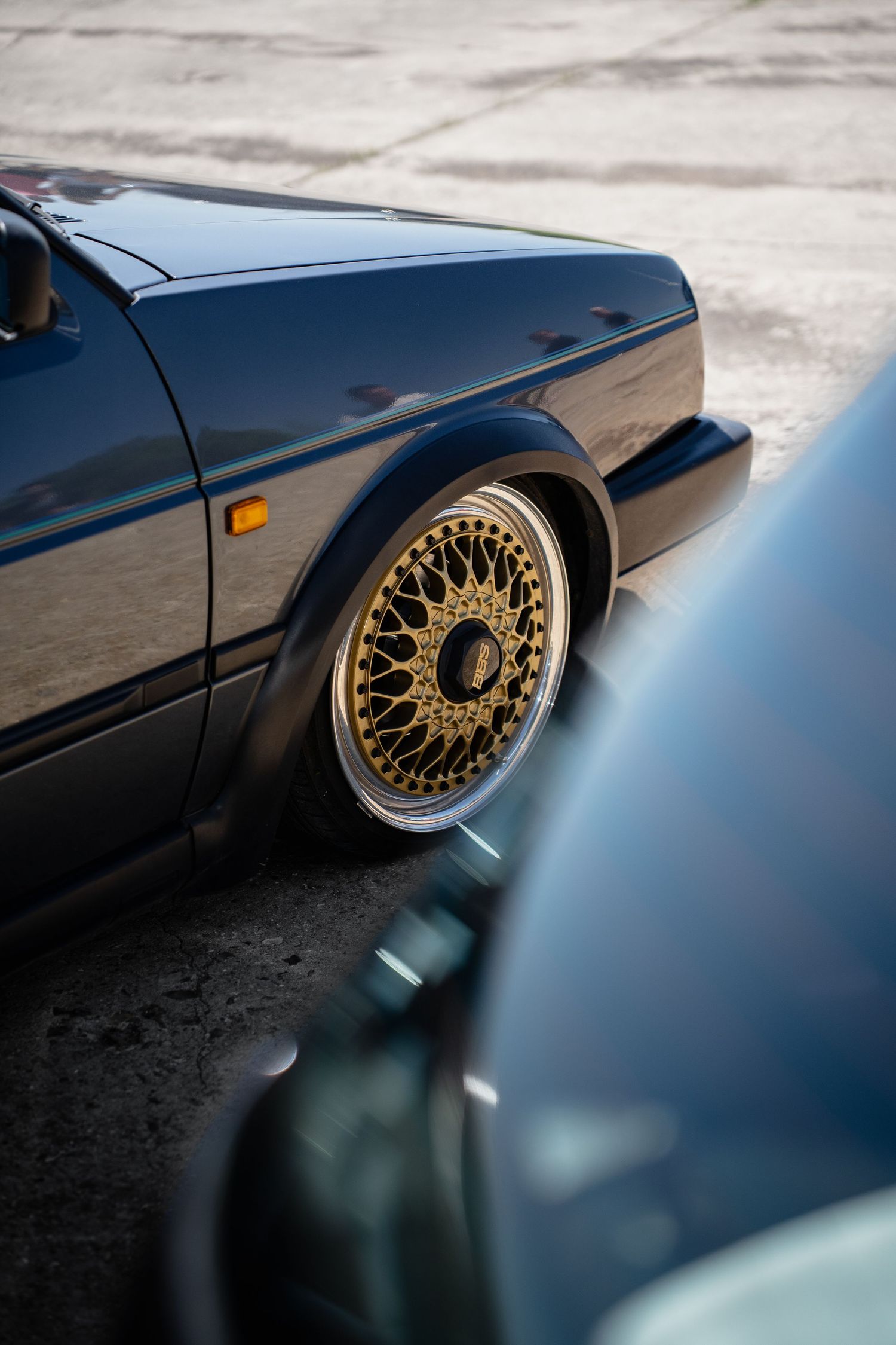 Dark blue classic car with gold BBS mesh wheels, lowered stance on concrete surface.