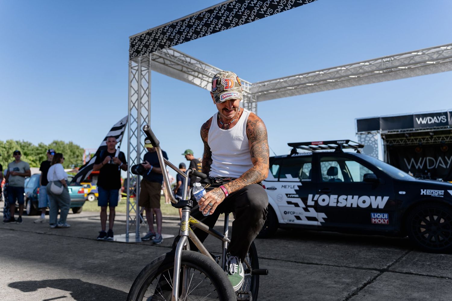 Man in white tank top riding a BMX bike at an outdoor event with a branded truck and metal arch in background.
