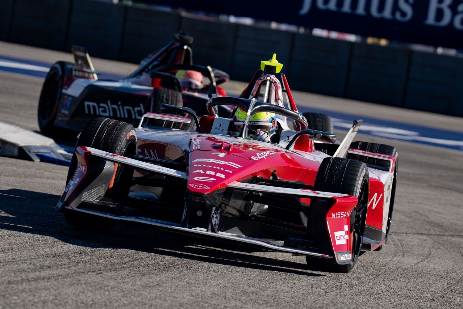 Two Formula E electric race cars compete on a street circuit, with a red car leading a Mahindra Racing car through a corner.