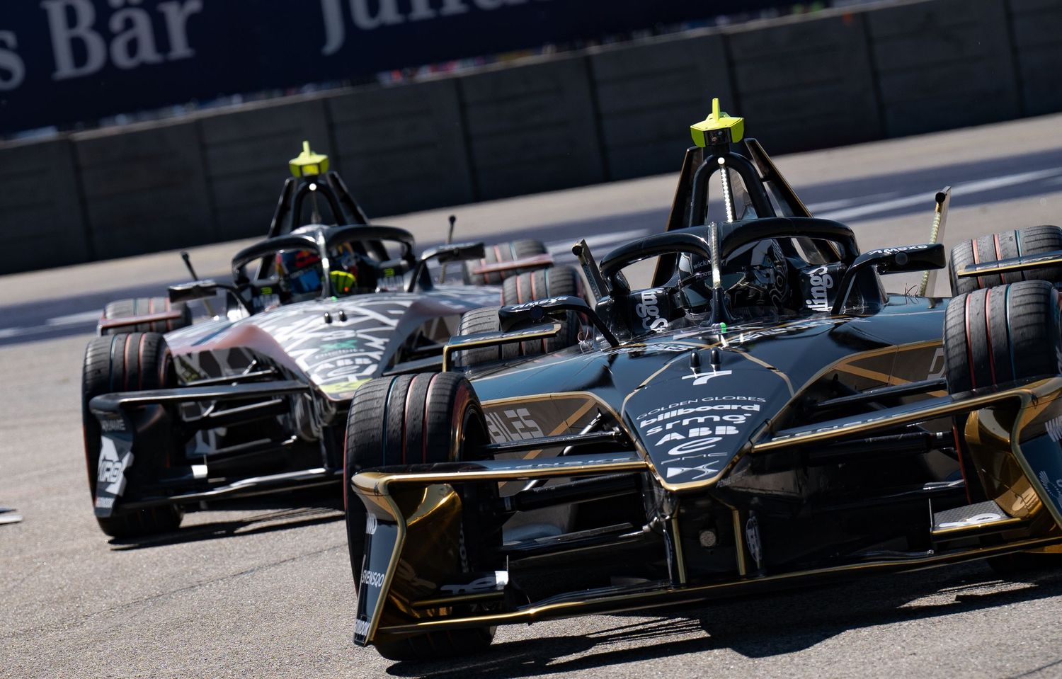 Two Formula E electric racing cars compete on a street circuit, with sponsors visible on barriers in the background.