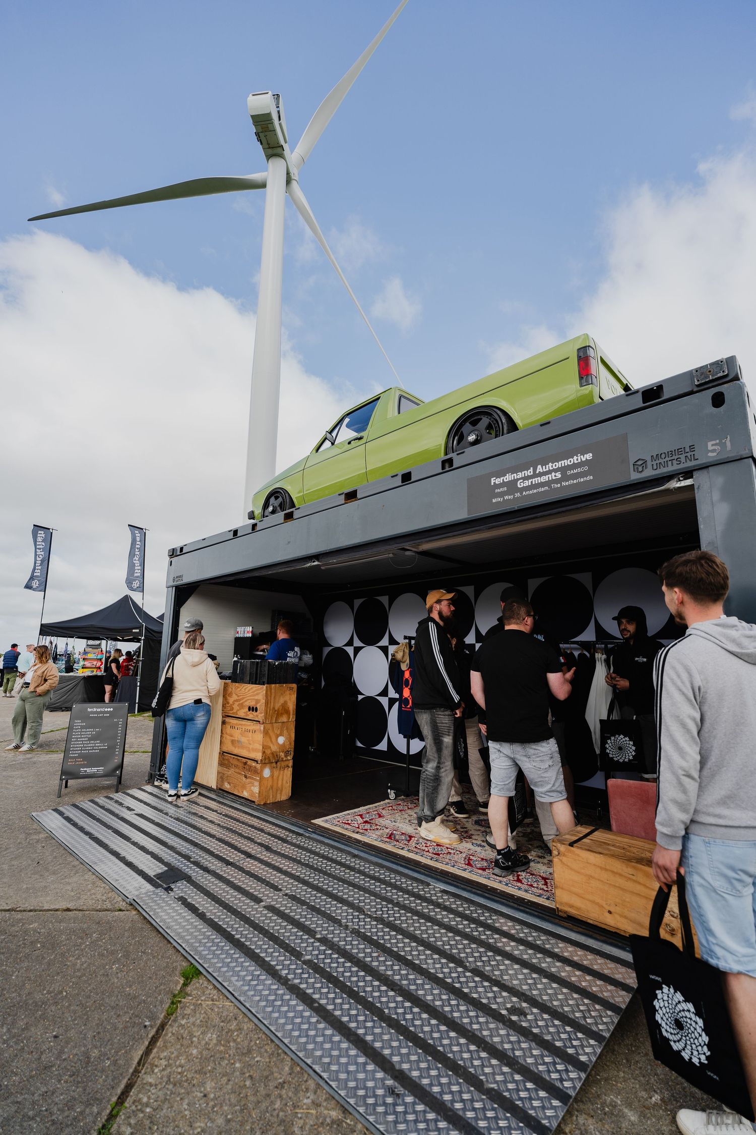 Green classic car displayed on top of a shipping container exhibit at an outdoor automotive event with a wind turbine backdrop.