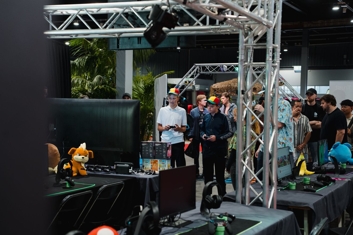 People gather around gaming displays and equipment at an indoor gaming convention with truss lighting rigs overhead.